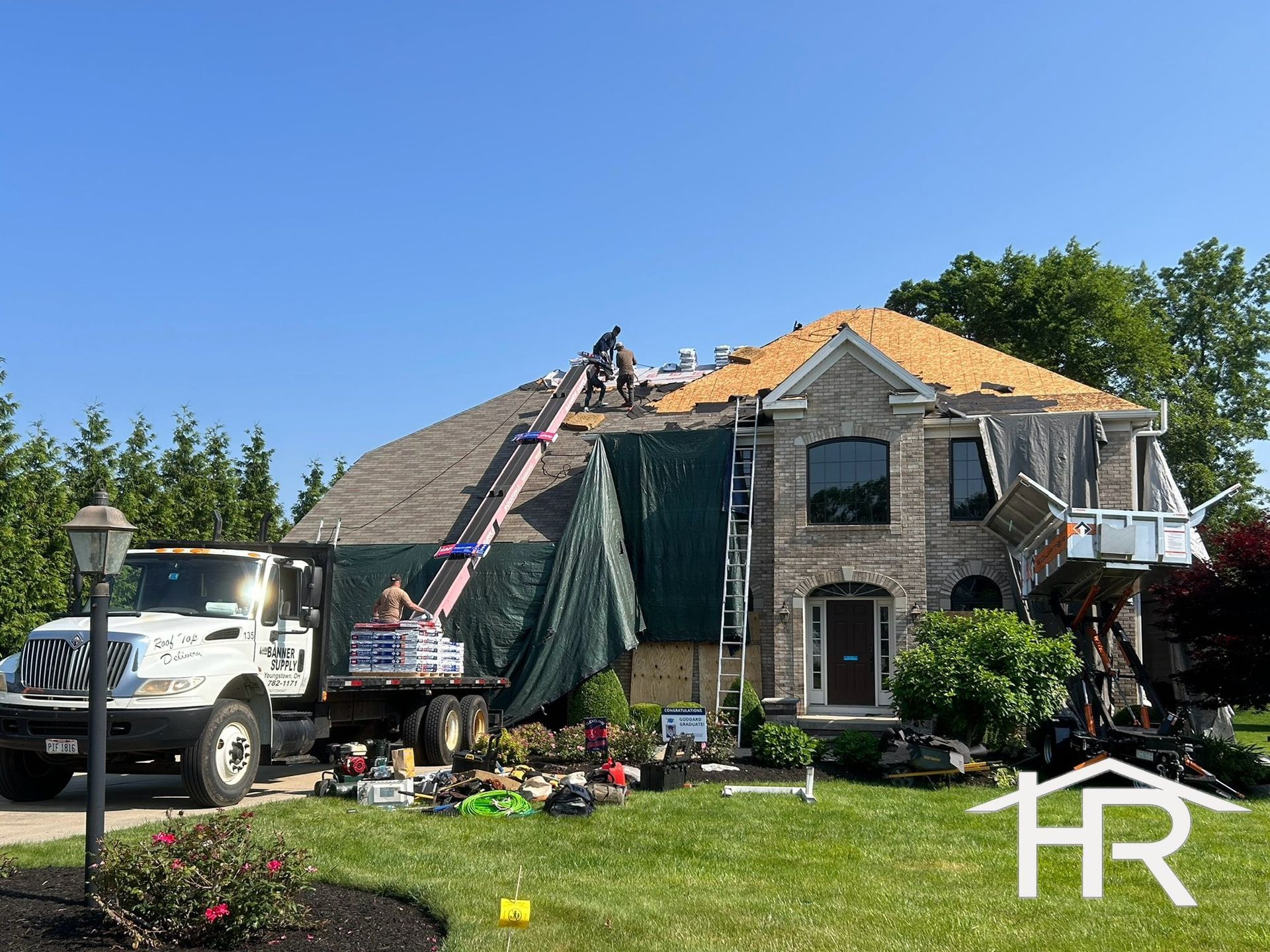 Roofers working on a house with a truck-mounted lift, green tarp, and debris on the lawn.