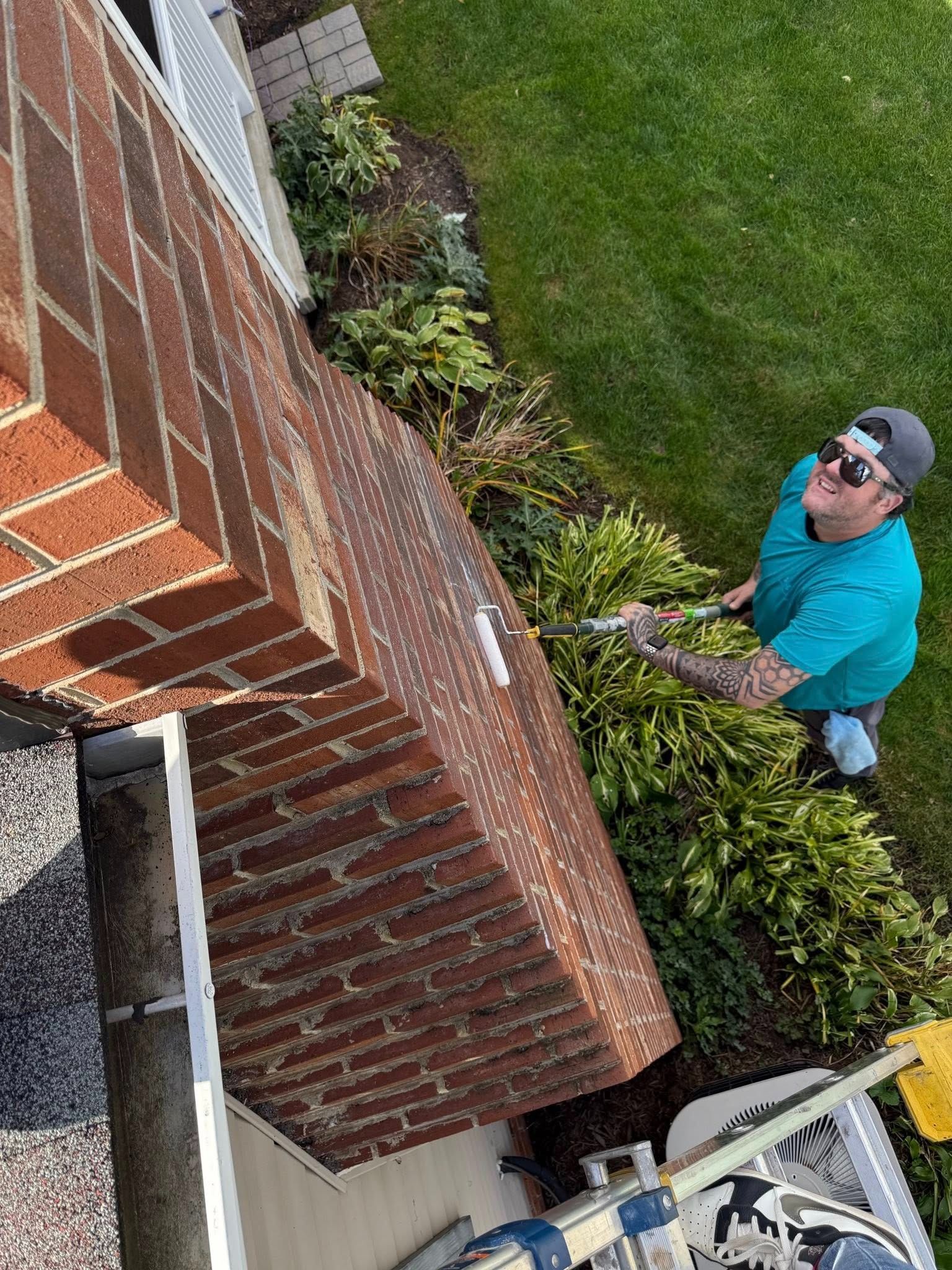 Person trimming bushes near a brick chimney, outside with green grass and white siding visible.
