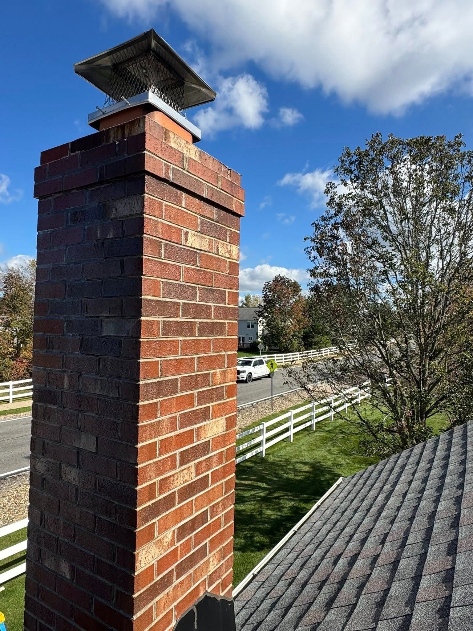Brick chimney with metal cap, on a roof with asphalt shingles, overlooking a street and sky.