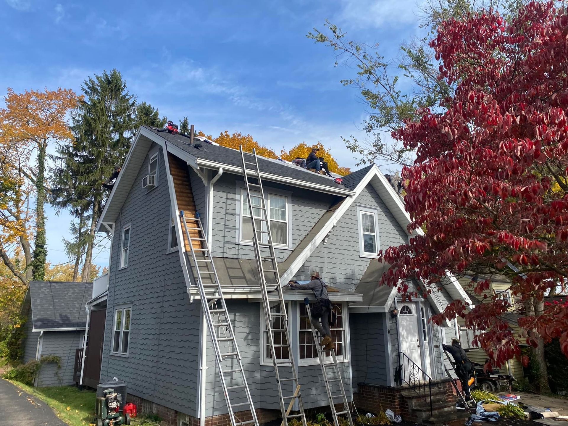 Workers on roof of gray house, installing new roofing on sunny day. Ladders, fall foliage present.