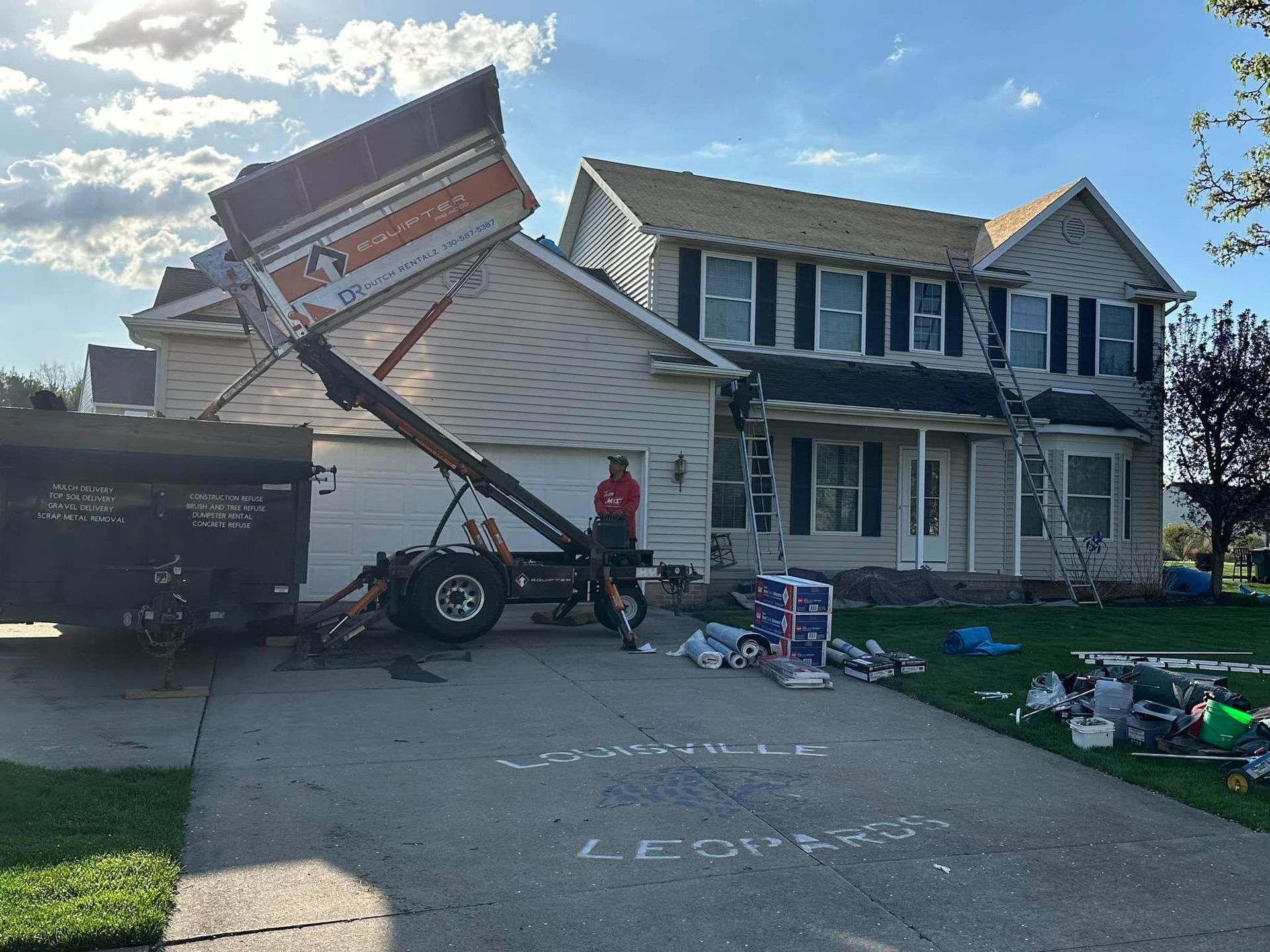 Dump truck raised in front of a house, man standing nearby, debris on the driveway.