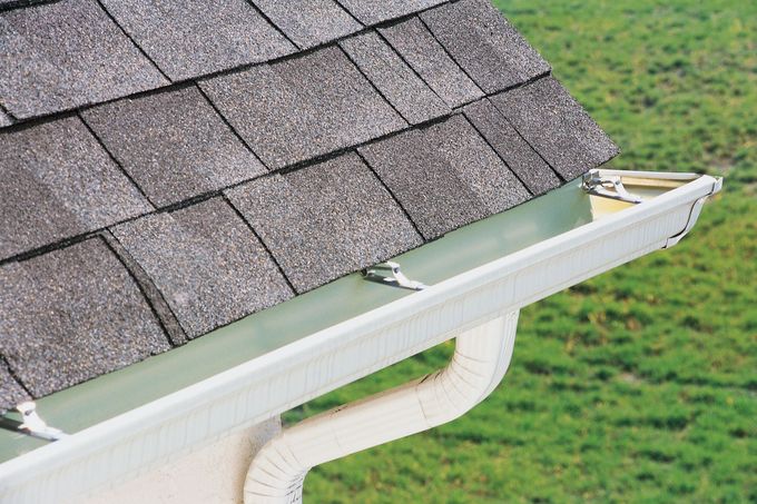 Gutter attached to a gray shingled roof, with white downspout against a green lawn.