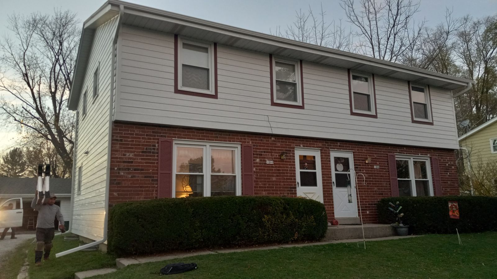 A man is standing in front of a brick house with a white siding.