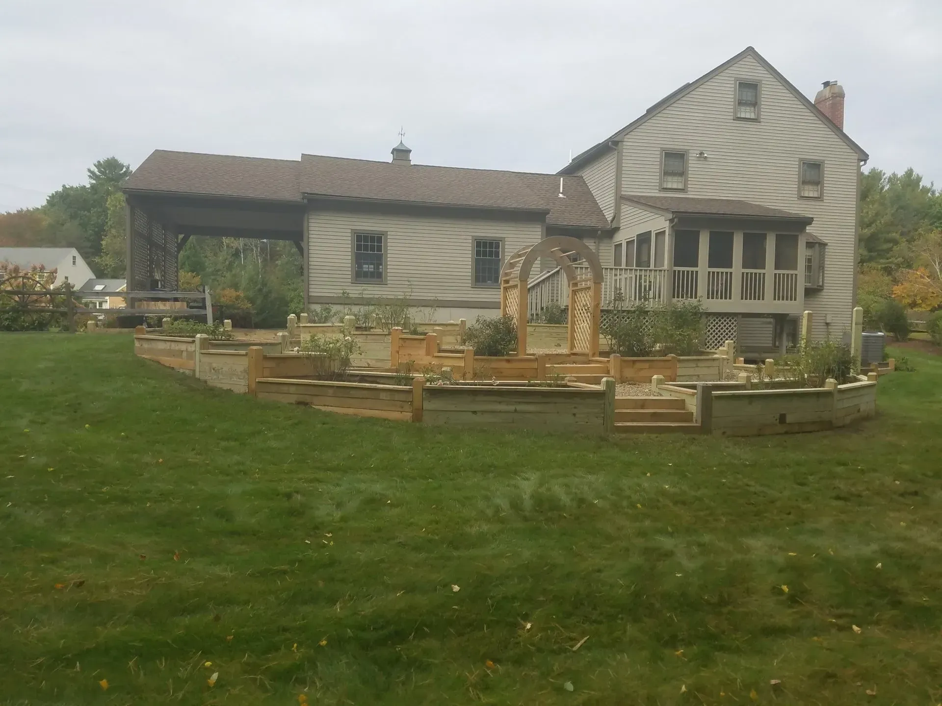 Rustic farmhouse with a wooden deck and fenced garden in a grassy yard under an overcast sky