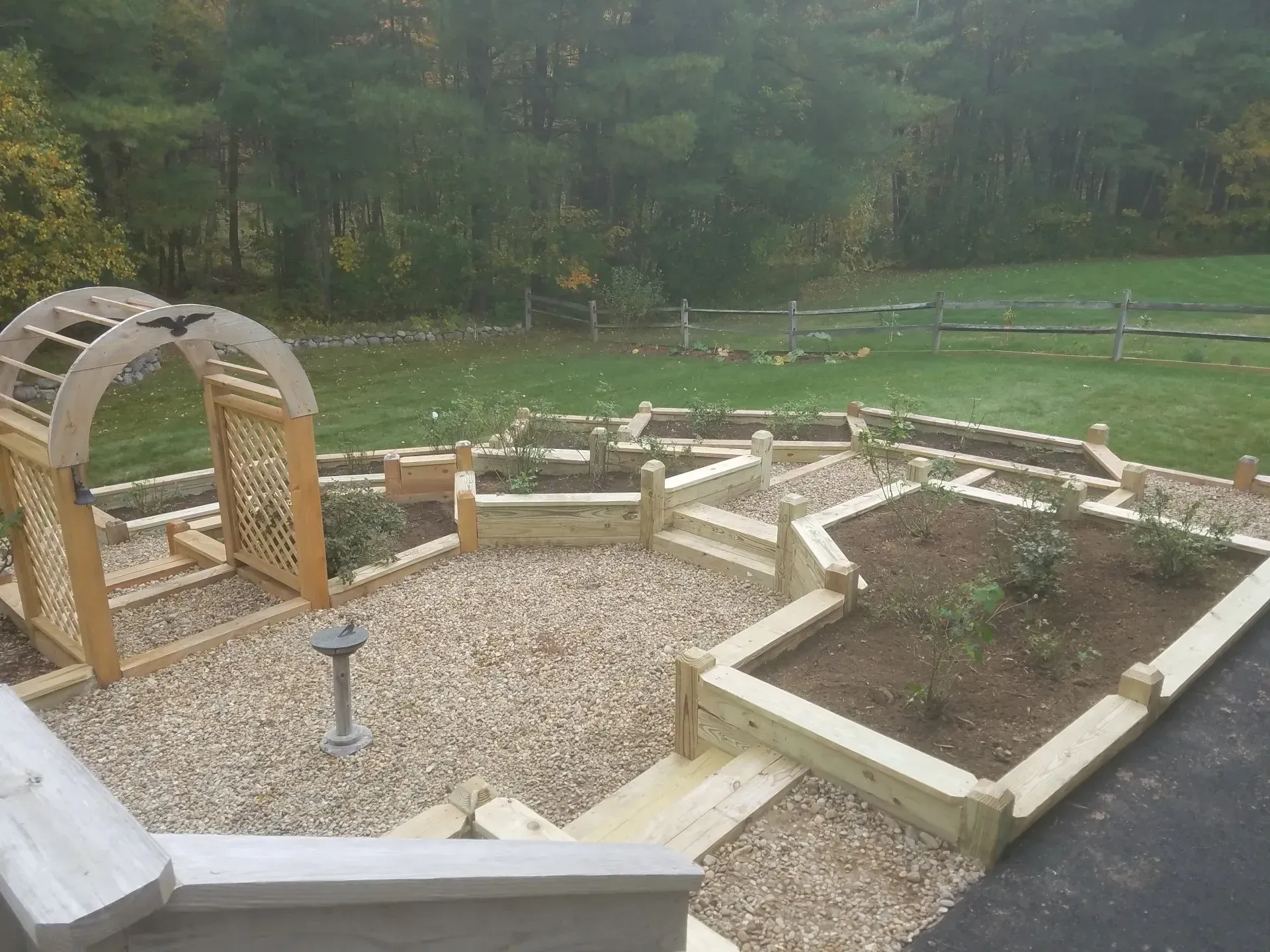 Raised garden beds and gravel paths beside a wooden arched trellis in a landscaped backyard.