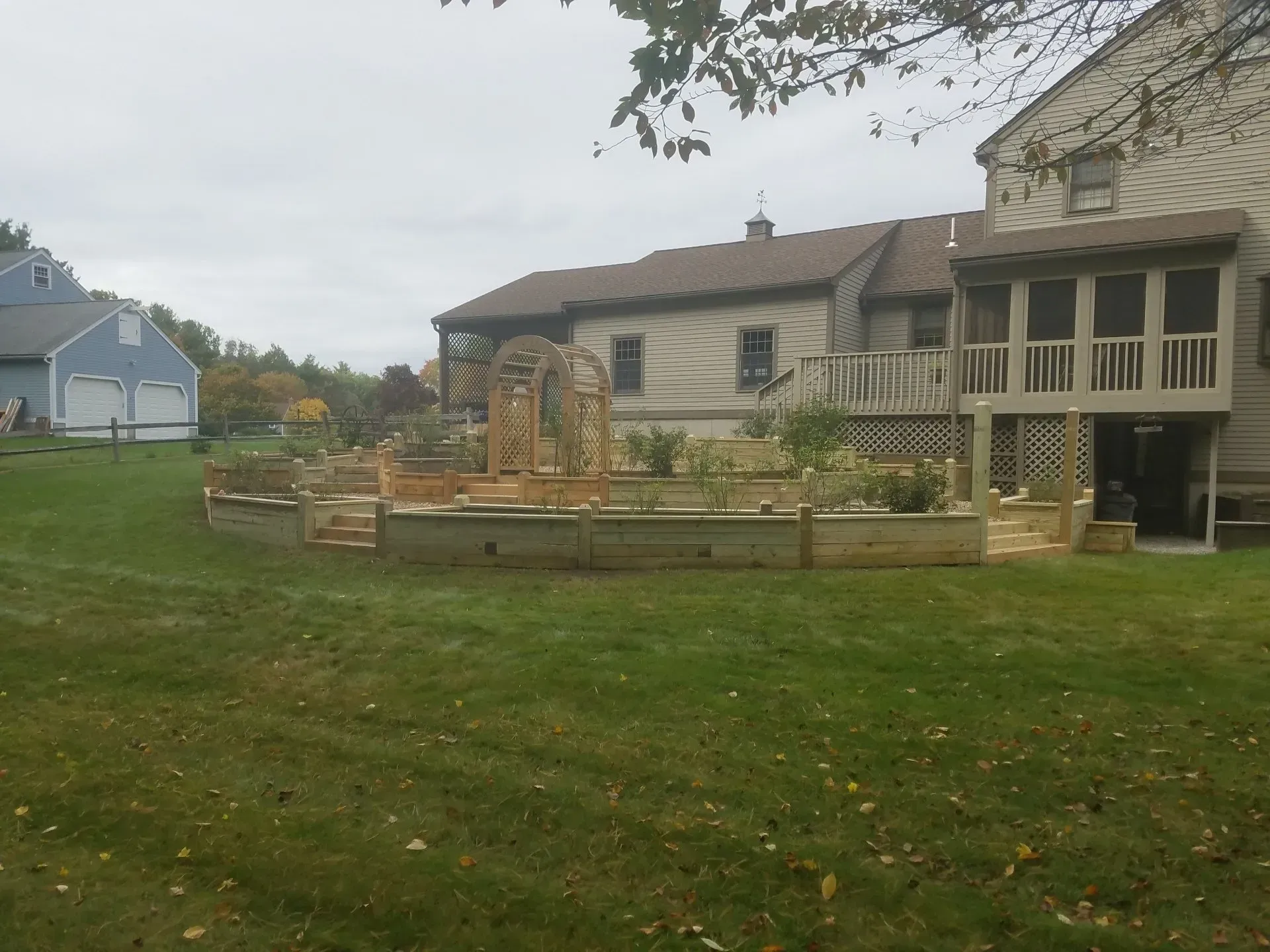 Backyard with raised garden beds, a small stone fountain, and a beige house with a porch under cloudy skies