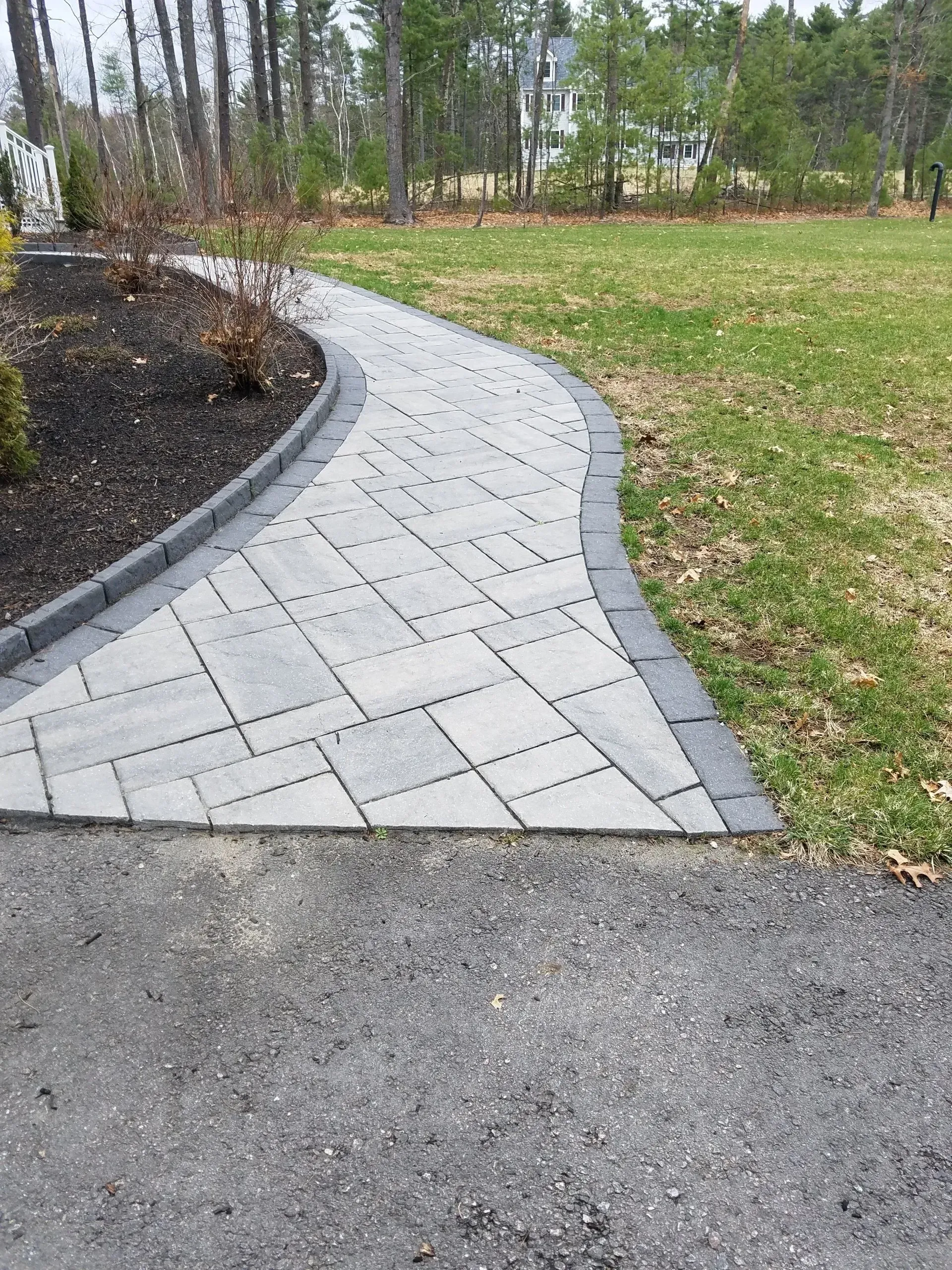 Curved gray paver walkway beside grass and a mulch bed in a wooded park area