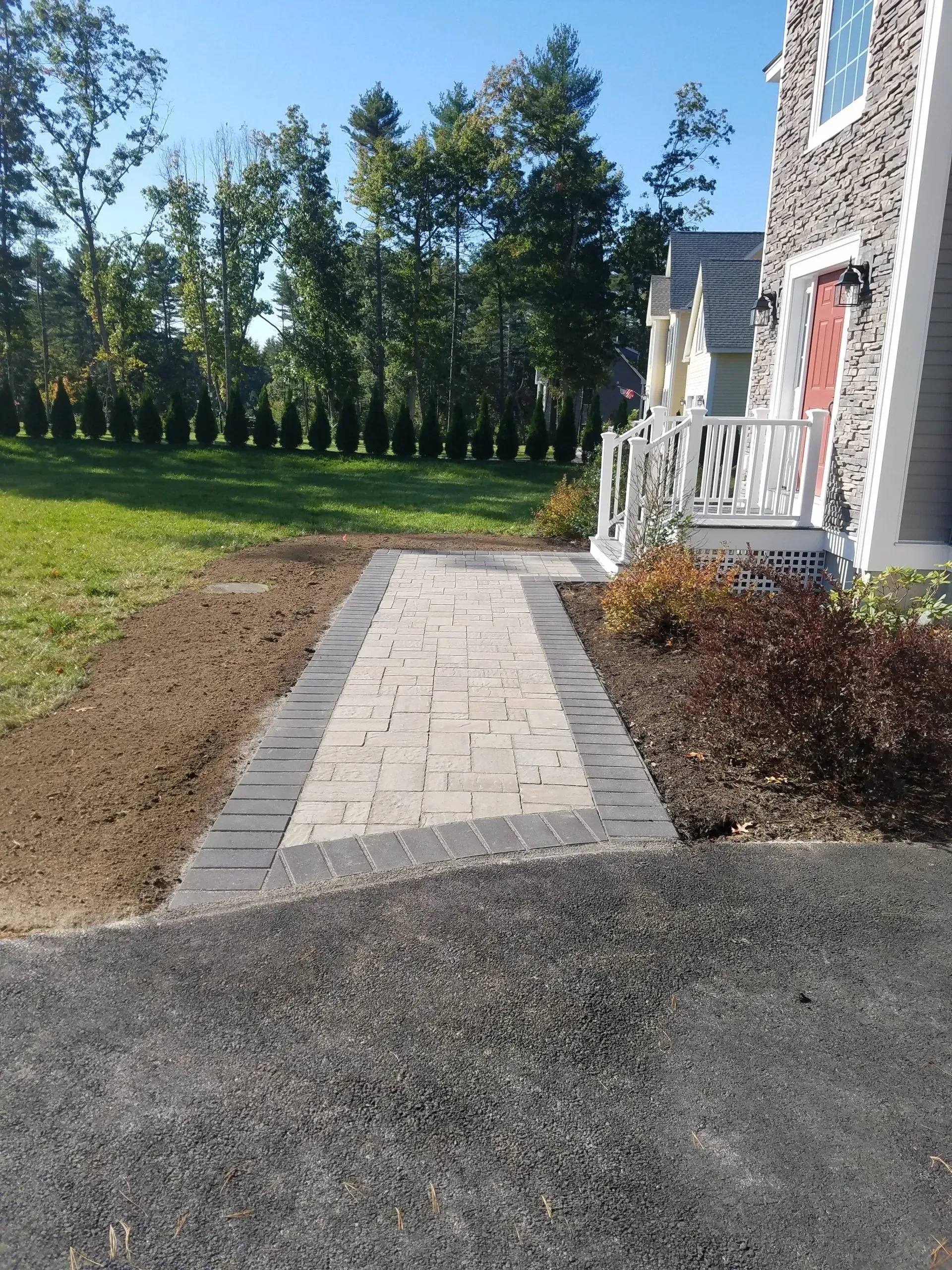 Paver walkway leading to a house entrance, bordered by grass, shrubs, and gravel driveway.