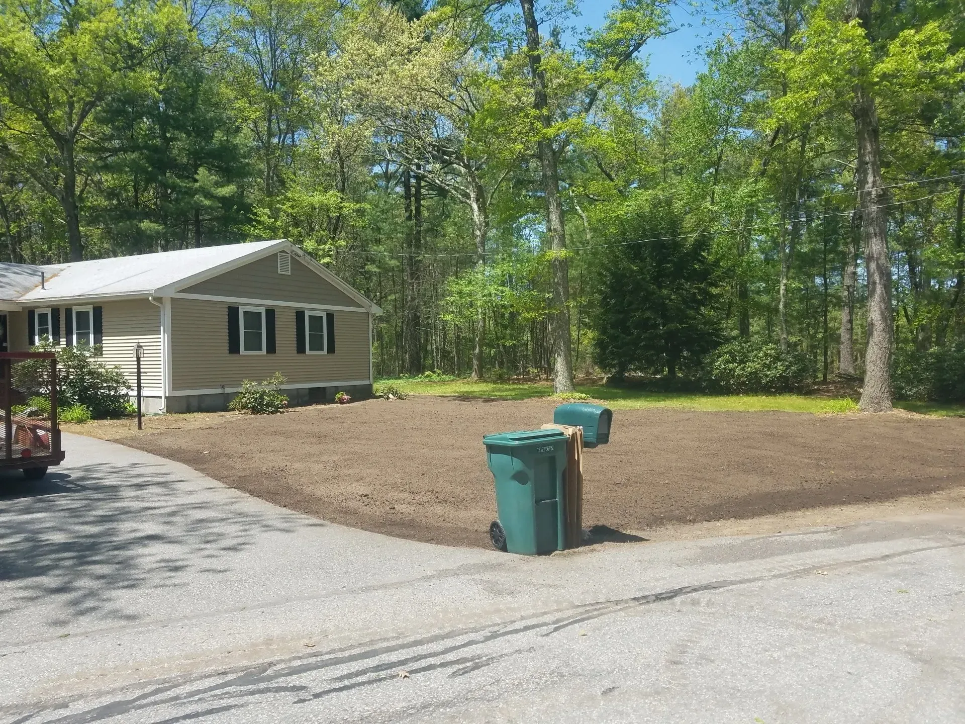 Small beige house by a gravel driveway with a dirt yard and green trees; two trash bins in front.