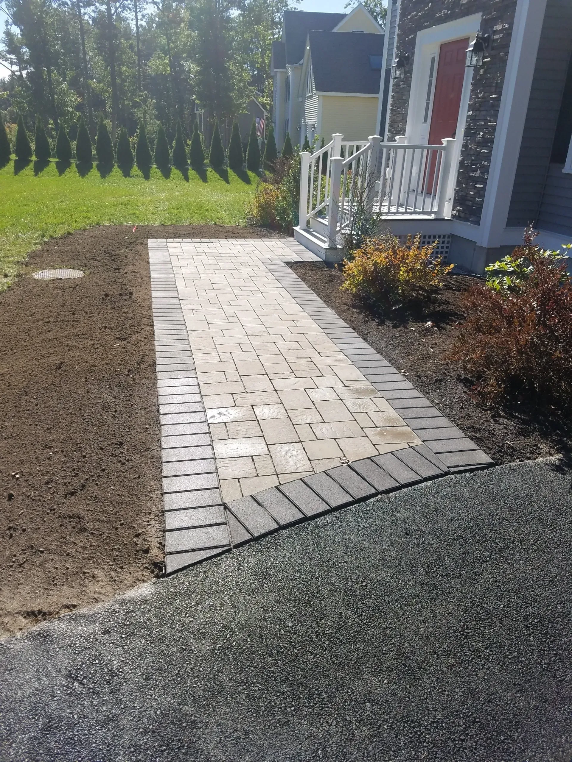 Brick walkway beside a house, bordered by fresh soil, gravel, and flower beds in a sunny yard