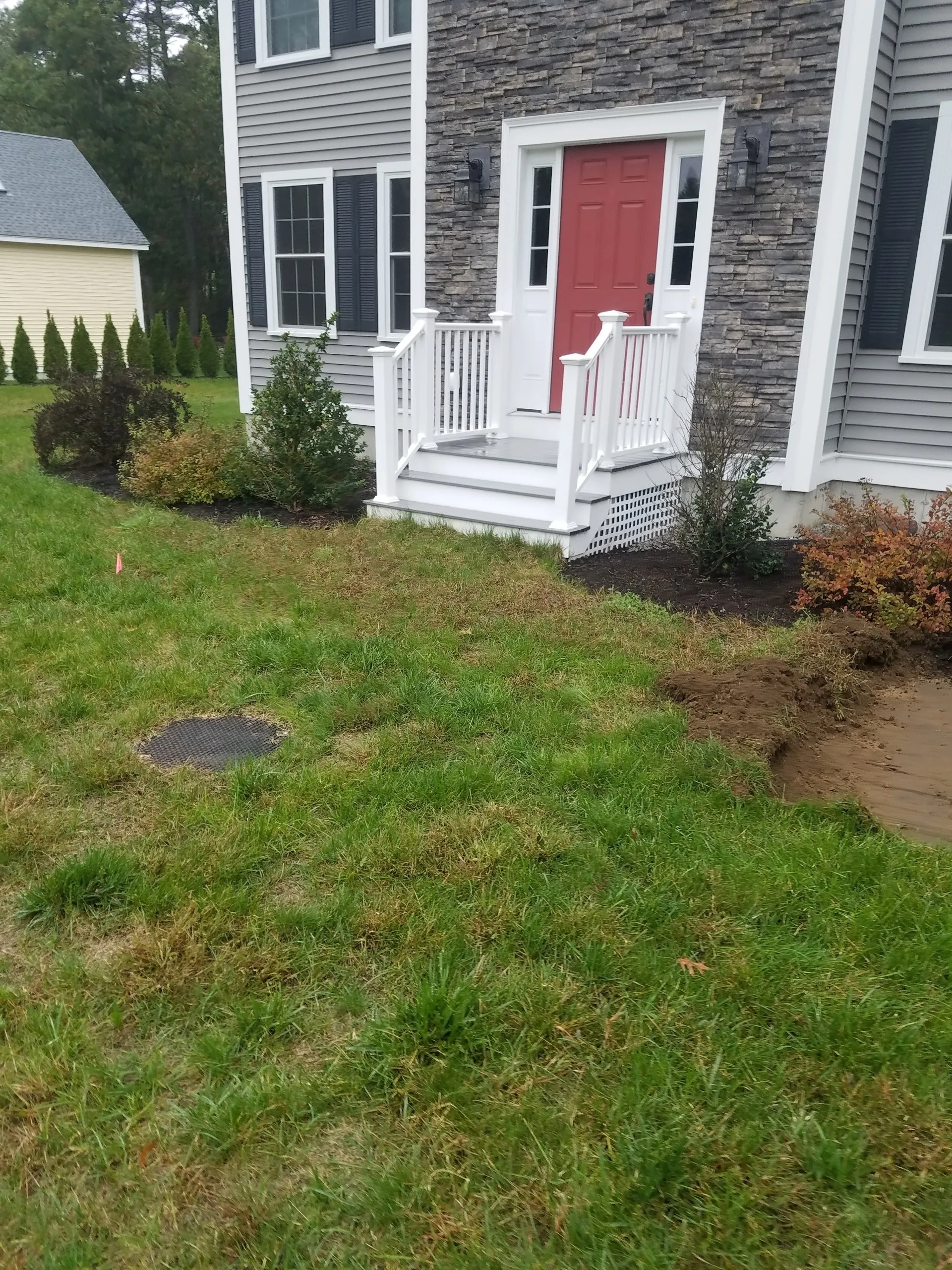 Front yard of a gray house with a red door and white porch, bordered by grass and shrubs.
