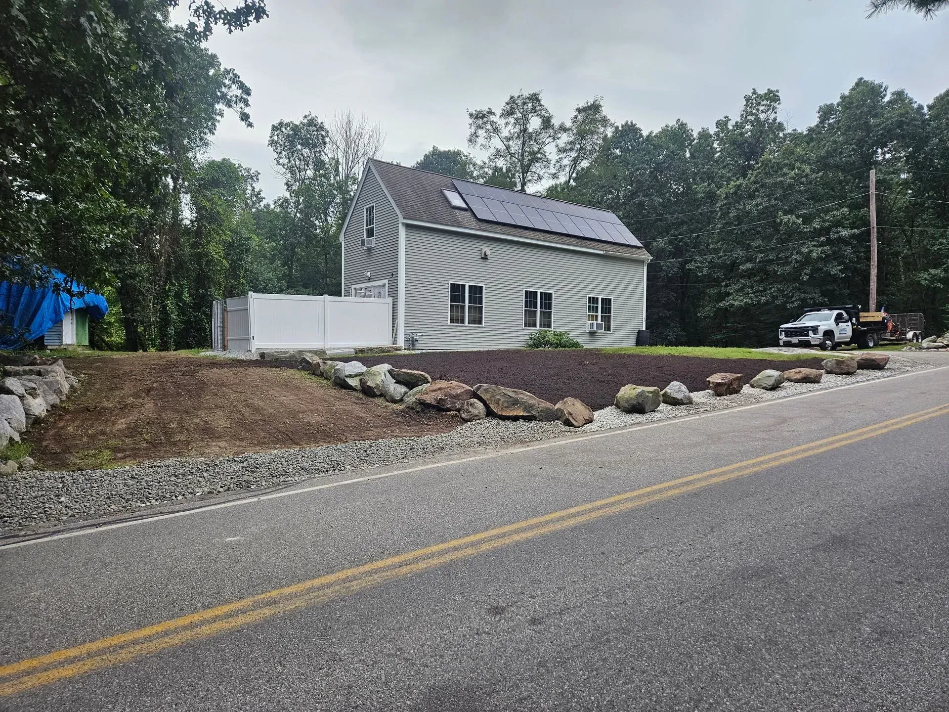 Small white house with a gray roof beside a road, with trees, rocks, and construction equipment nearby