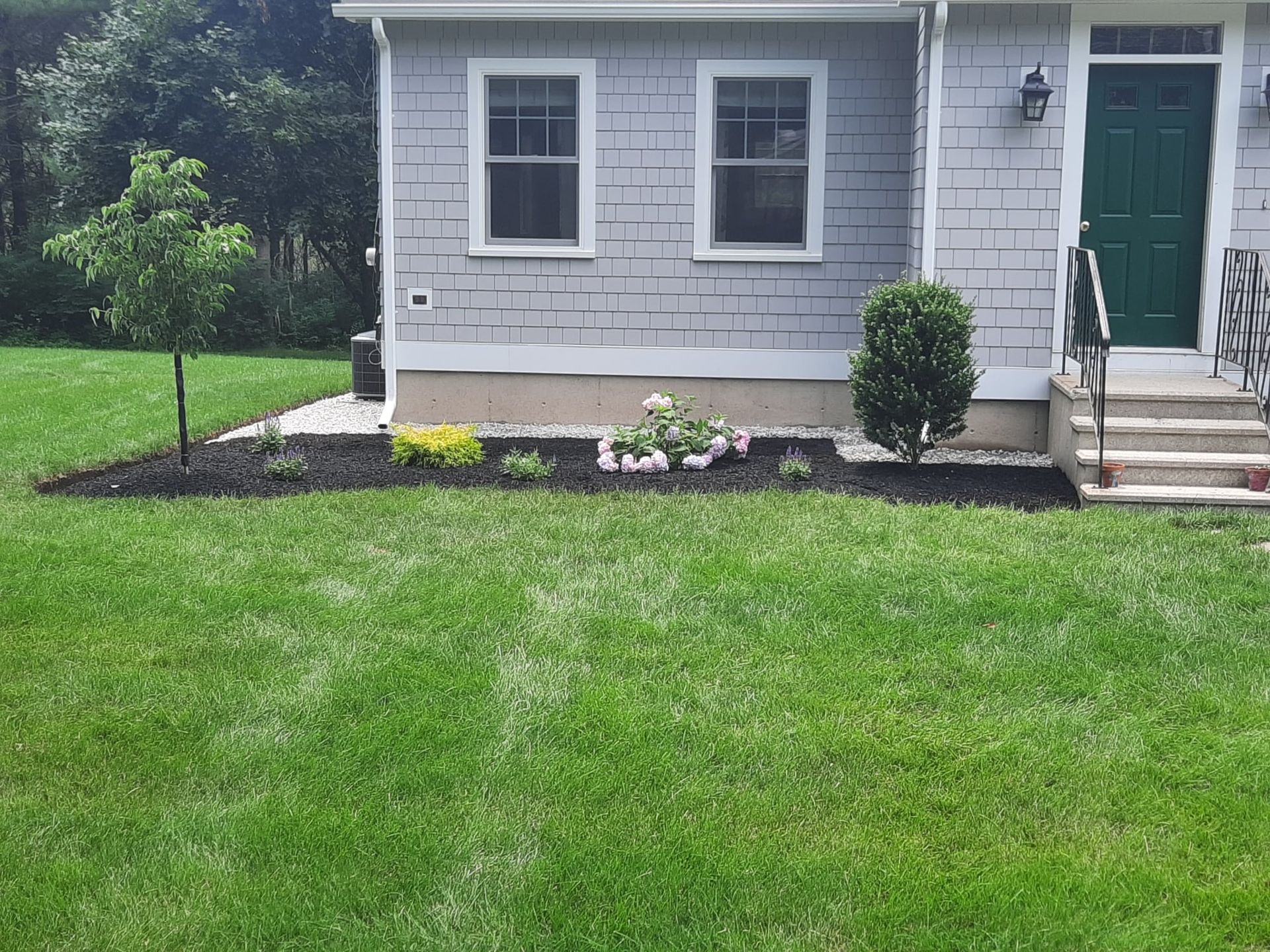 Front lawn and gray house exterior with green door, windows, and flower bed by the steps