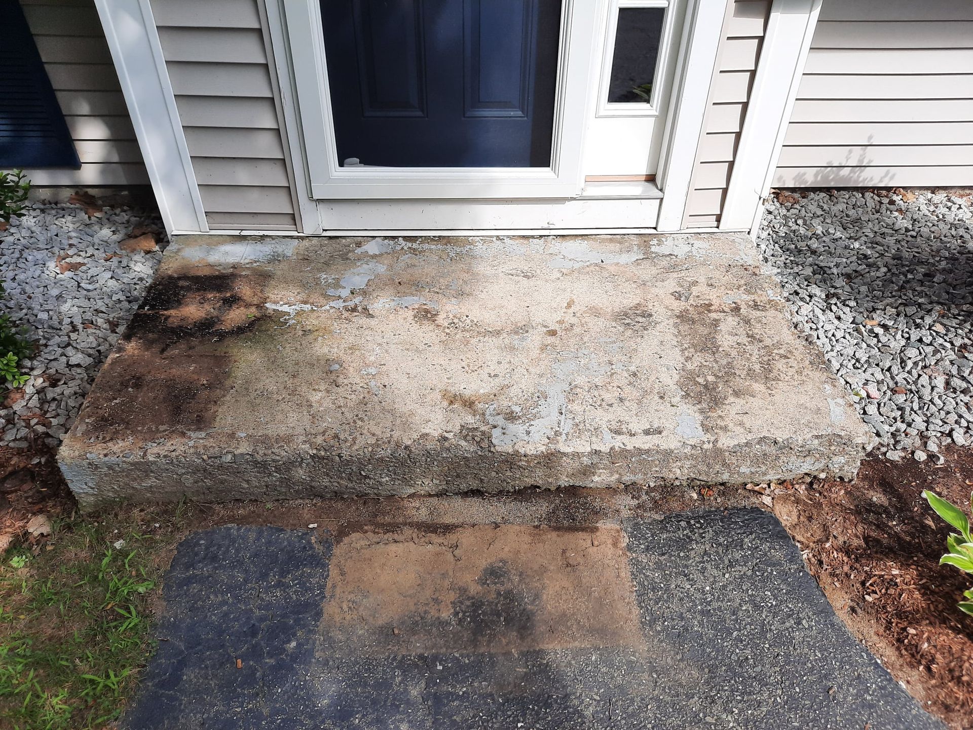 Weathered concrete front step with a small doormat, bordered by gray siding and gravel.