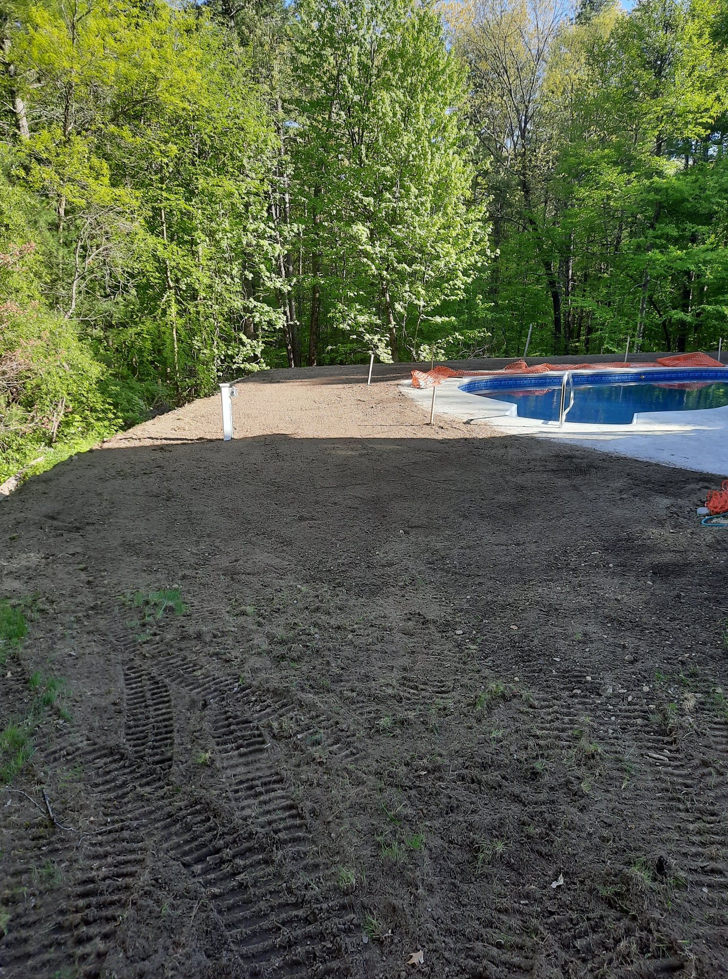 Freshly graded dirt beside a blue above-ground pool in a wooded backyard