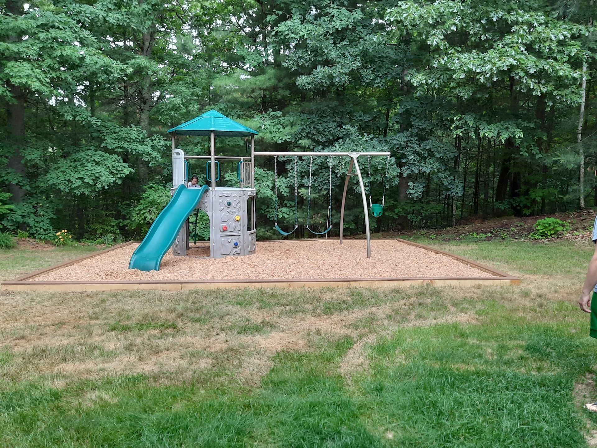 Playground with blue slide, swing set, and climbing tower on a wood-chip surface, surrounded by trees