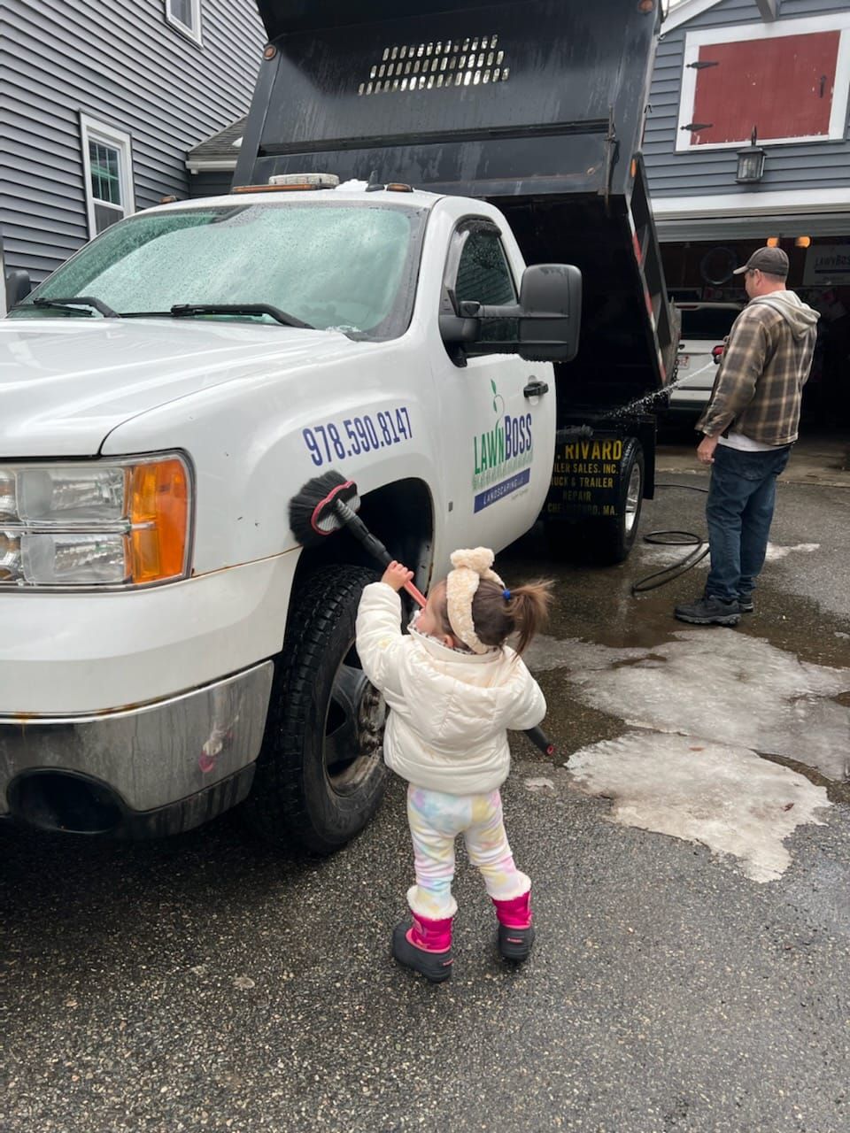 Child in winter clothes reaching toward a white utility truck with an open rear liftgate in a driveway.