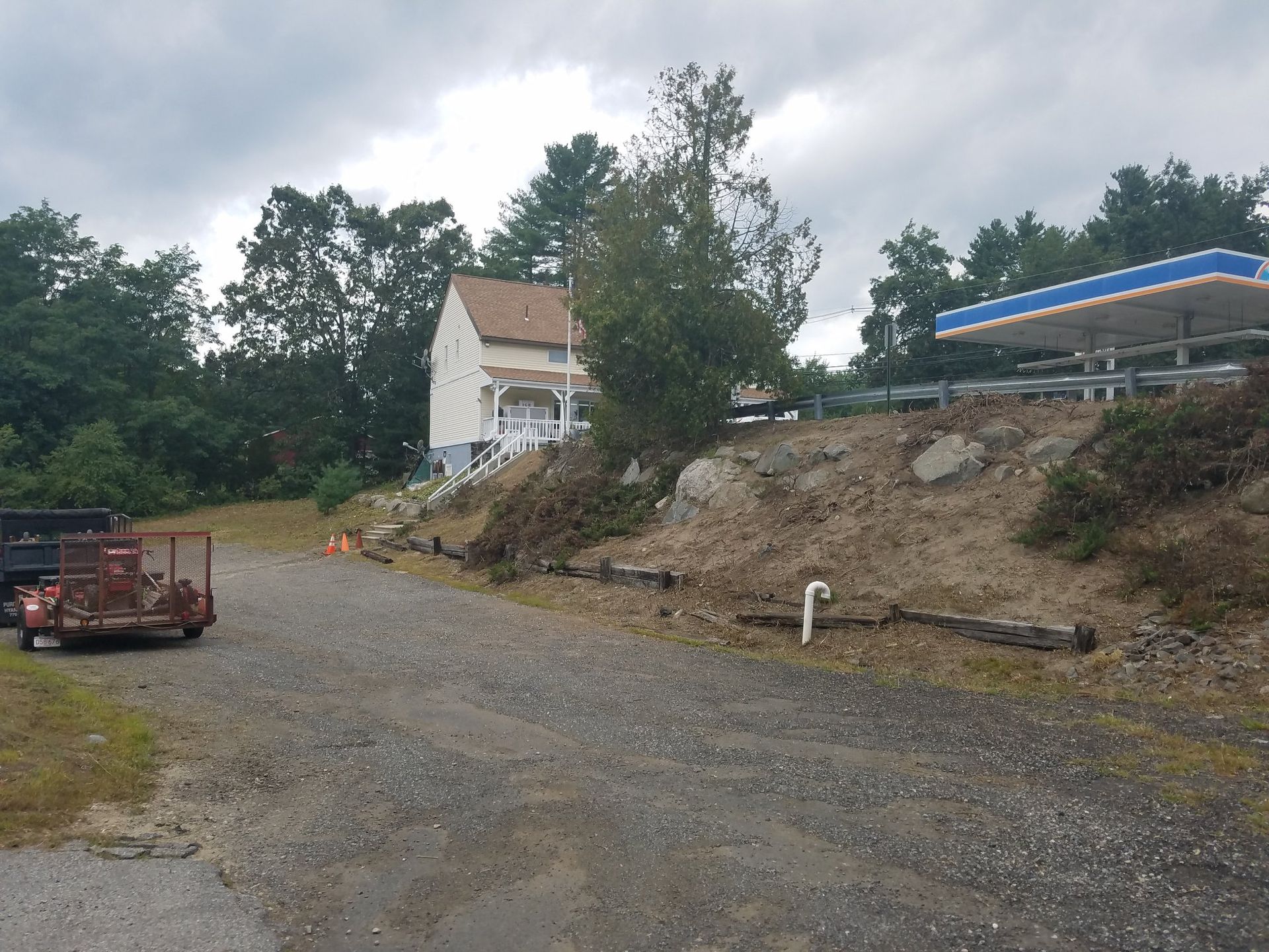 Gravel lot with a white house on a slope and a gas station canopy beside trees under cloudy skies