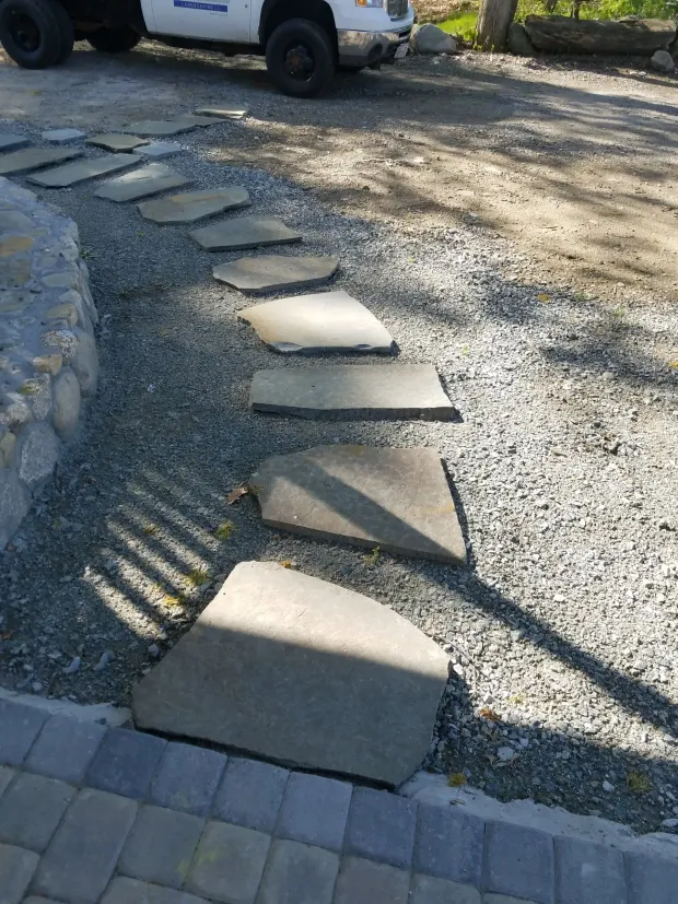 Stepping stones across a gravel path beside a stone wall, with a parked truck in the background.