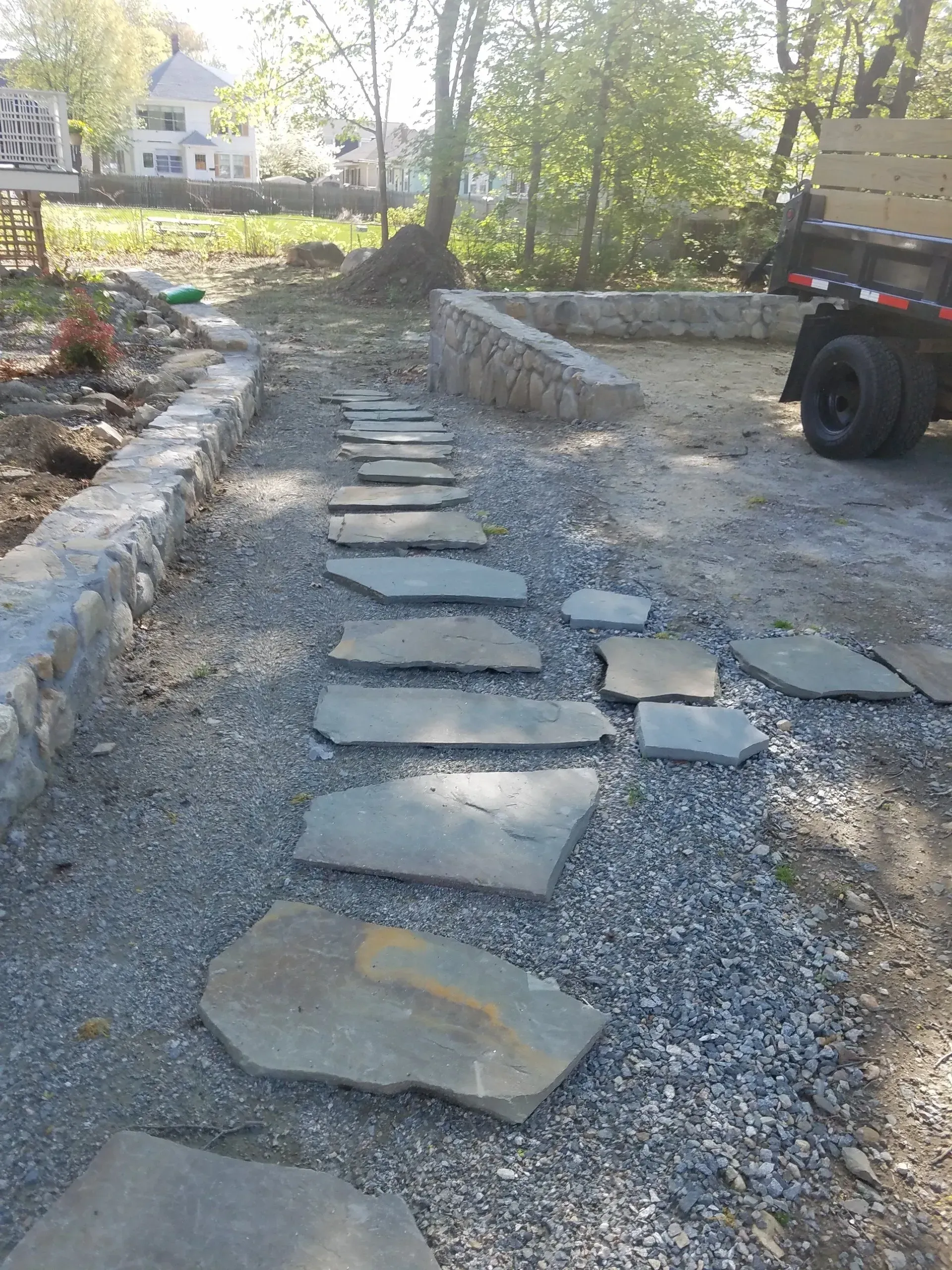 Gravel path with uneven stepping stones beside a low stone wall in a yard, with a vehicle on the right.