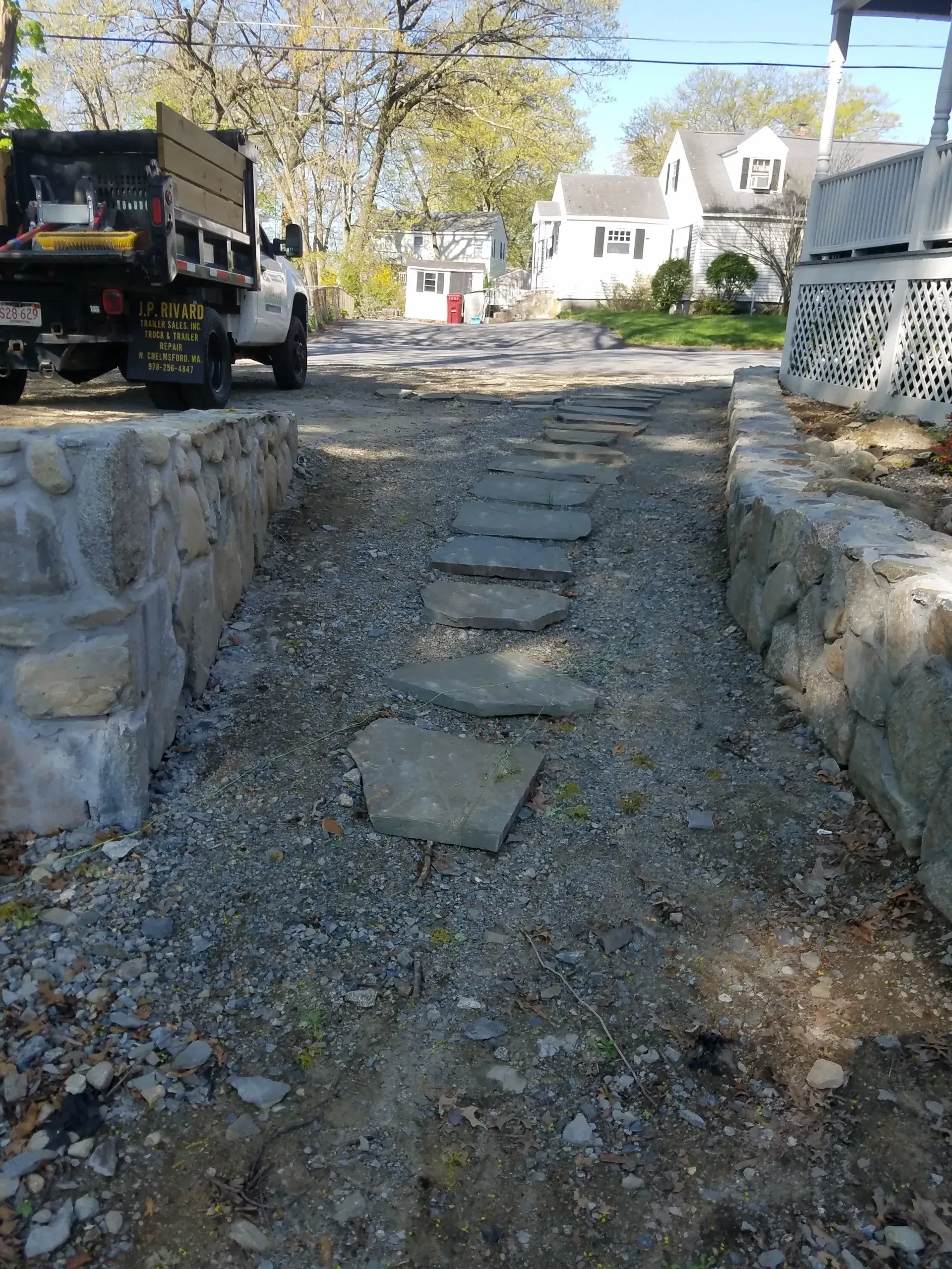 Stone steps leading up a gravel path between stone walls, with a truck and houses in the background.