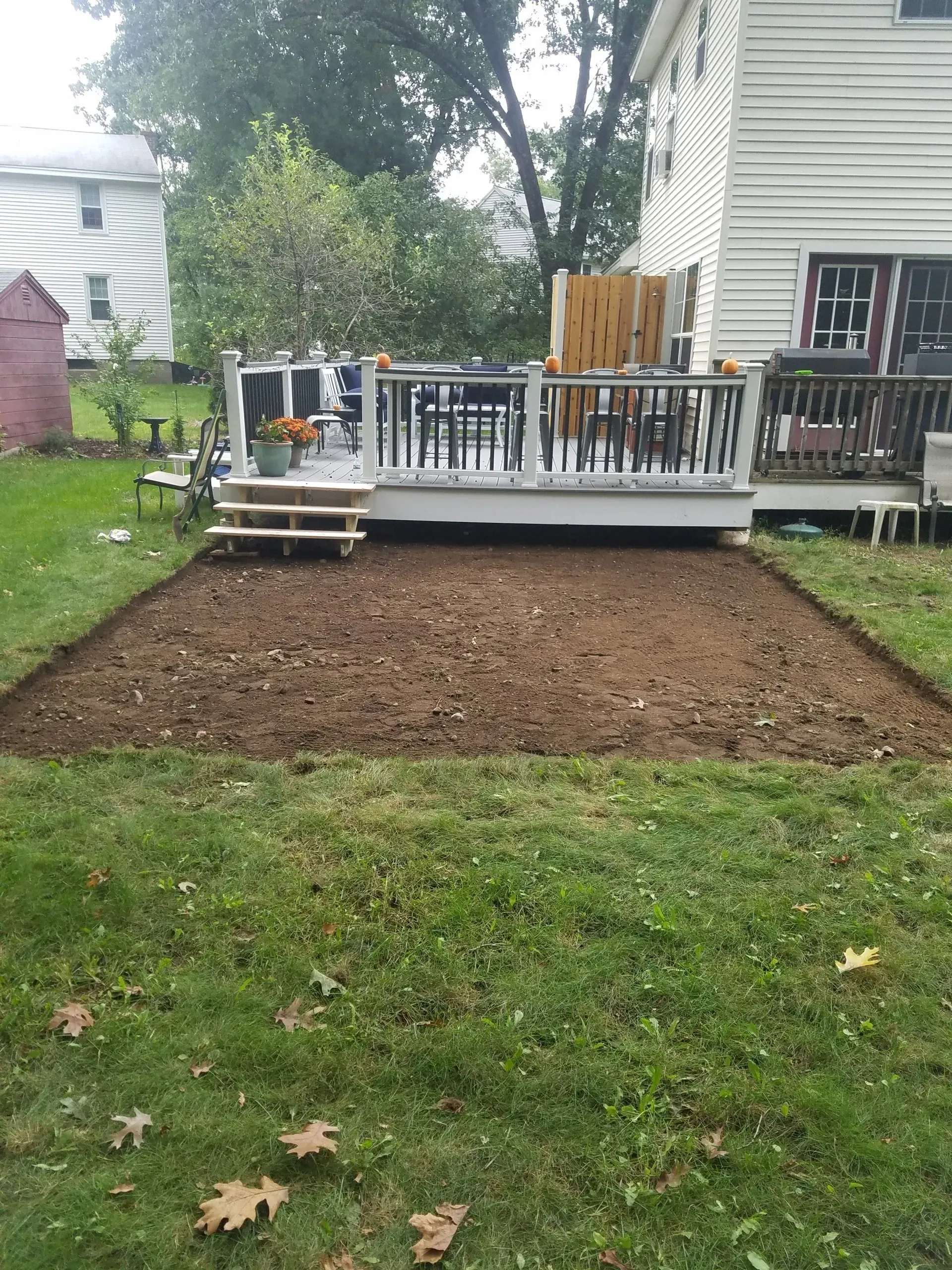 Freshly tilled soil bed beside a white house and wooden deck in a backyard.