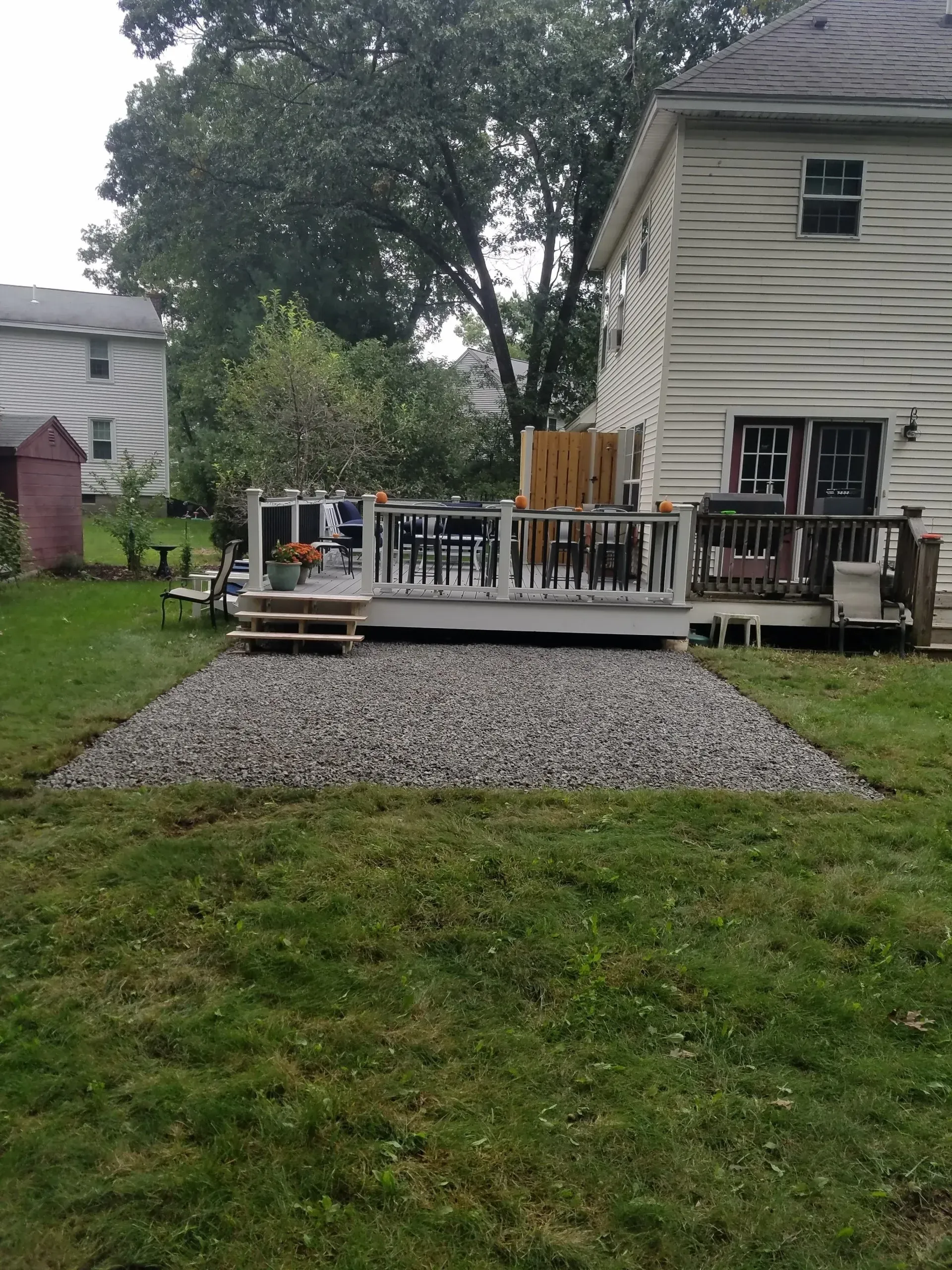 Backyard deck with white railing, steps, gravel path, and house on the right beside a tree-lined yard
