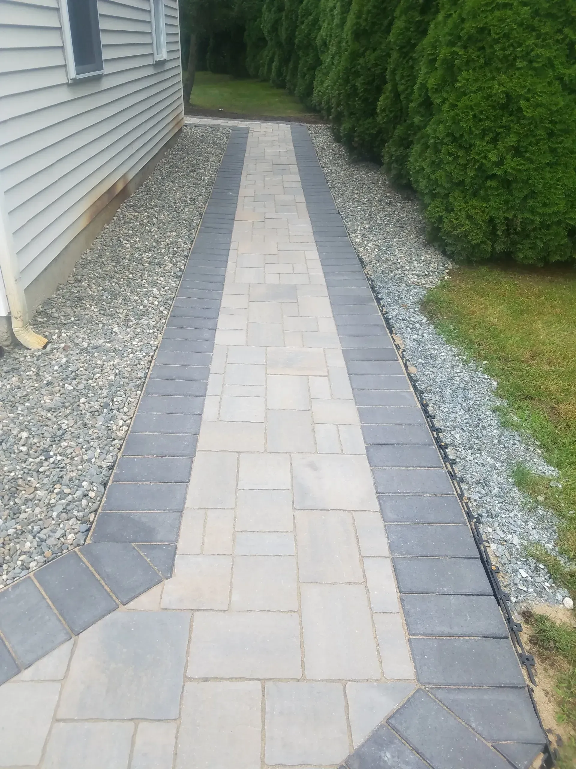 Narrow paver walkway beside a house, bordered by gray gravel and green hedges.