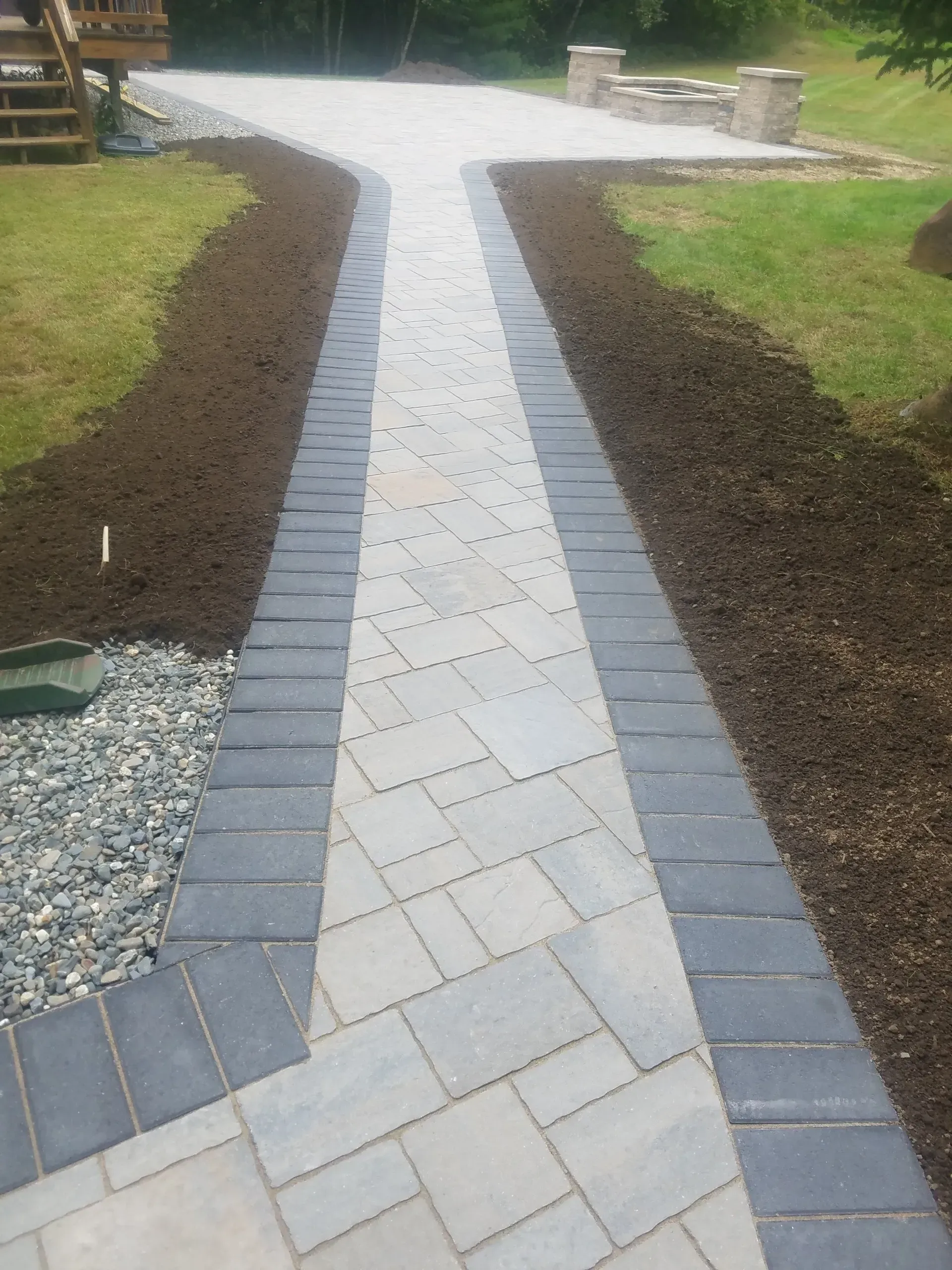 Curved stone garden path bordered by dark pavers, mulch, grass, and gravel leading to a patio area