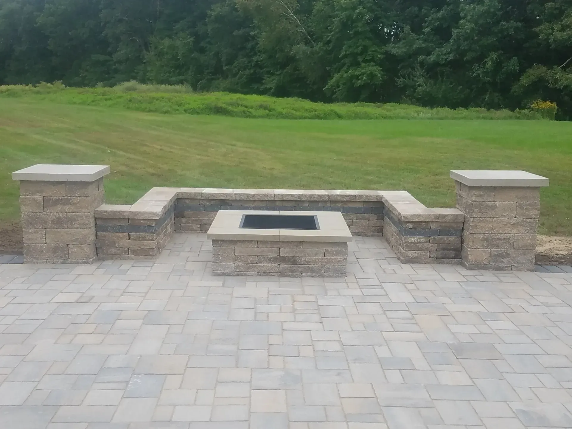 Stone patio with a low fire pit wall overlooking a grassy lawn and trees.