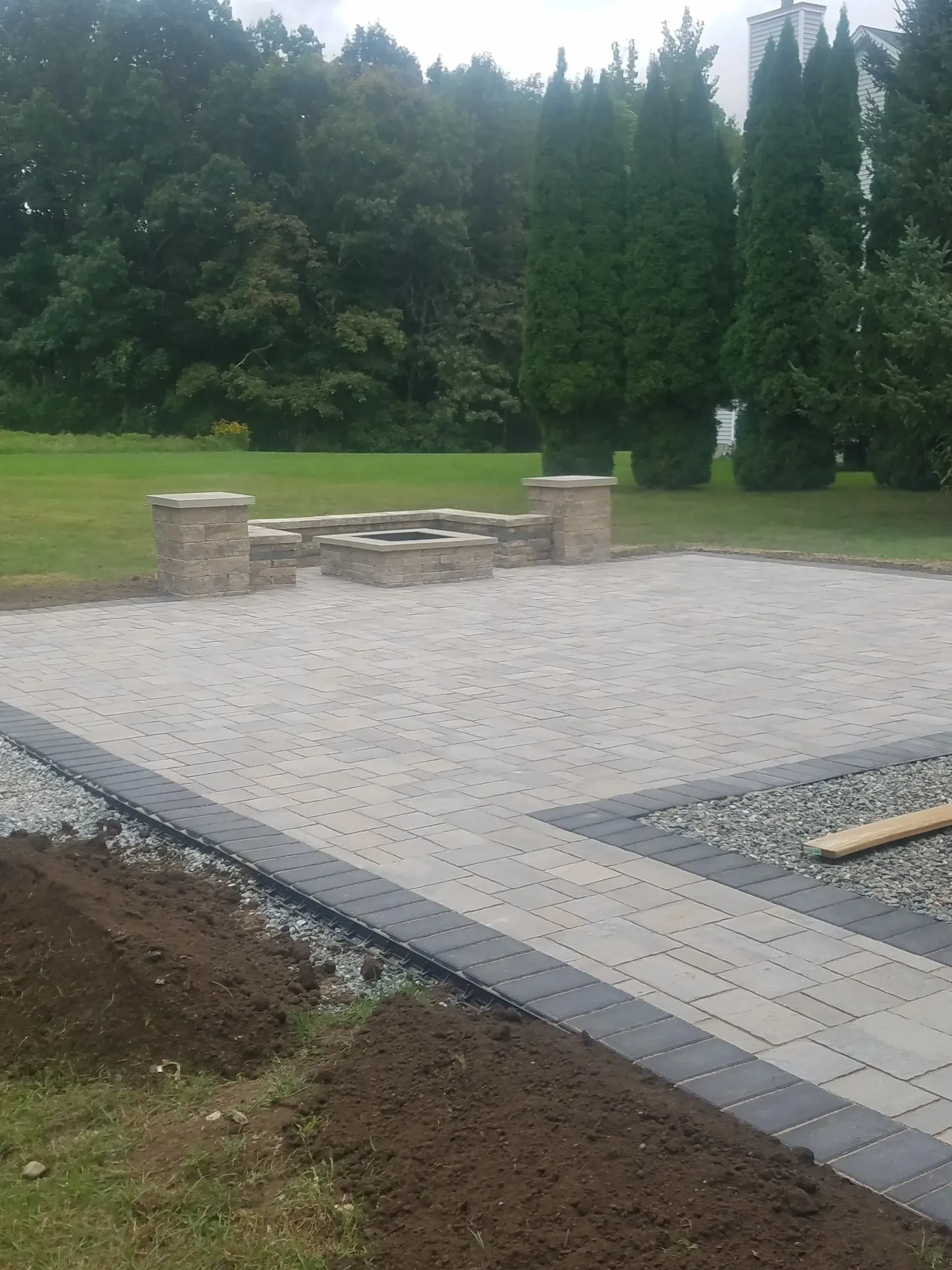 Paved patio under construction beside a lawn, with stone seating and trees in the background.