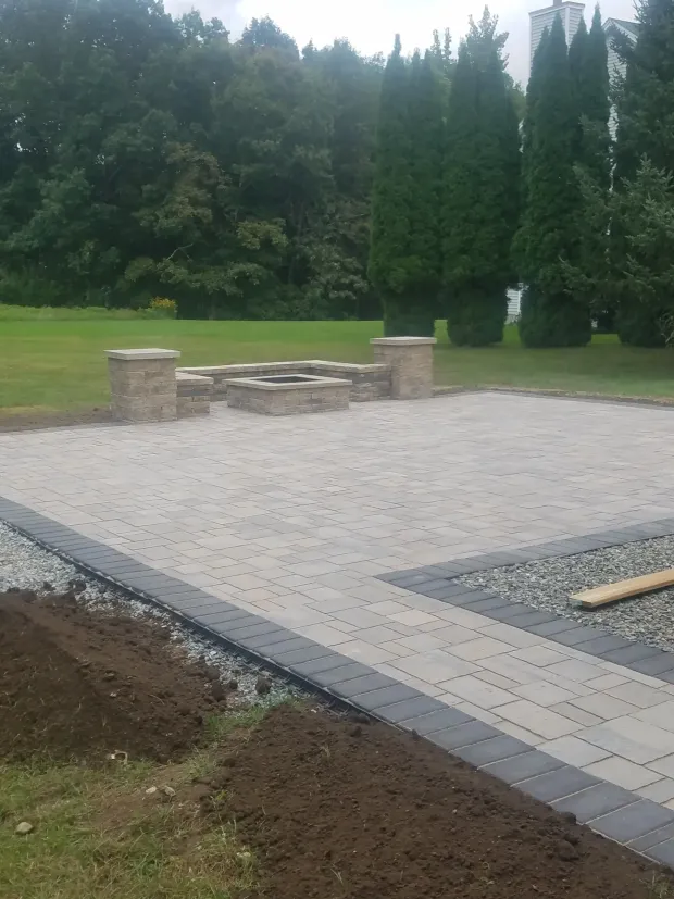 Outdoor patio with stone pavers, built-in seating, and a grassy yard bordered by tall trees.