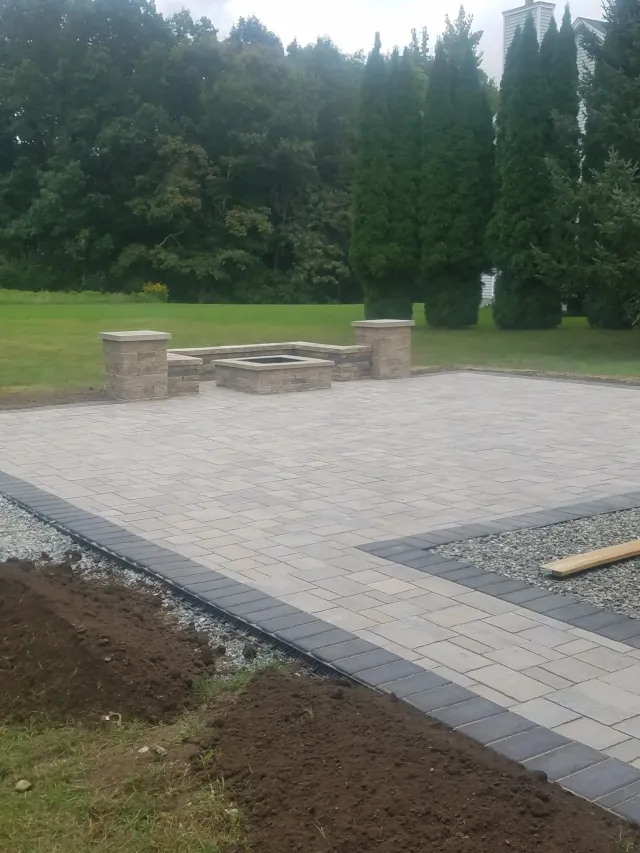 Outdoor patio with stone pavers, built-in seating, and a grassy yard bordered by tall trees.