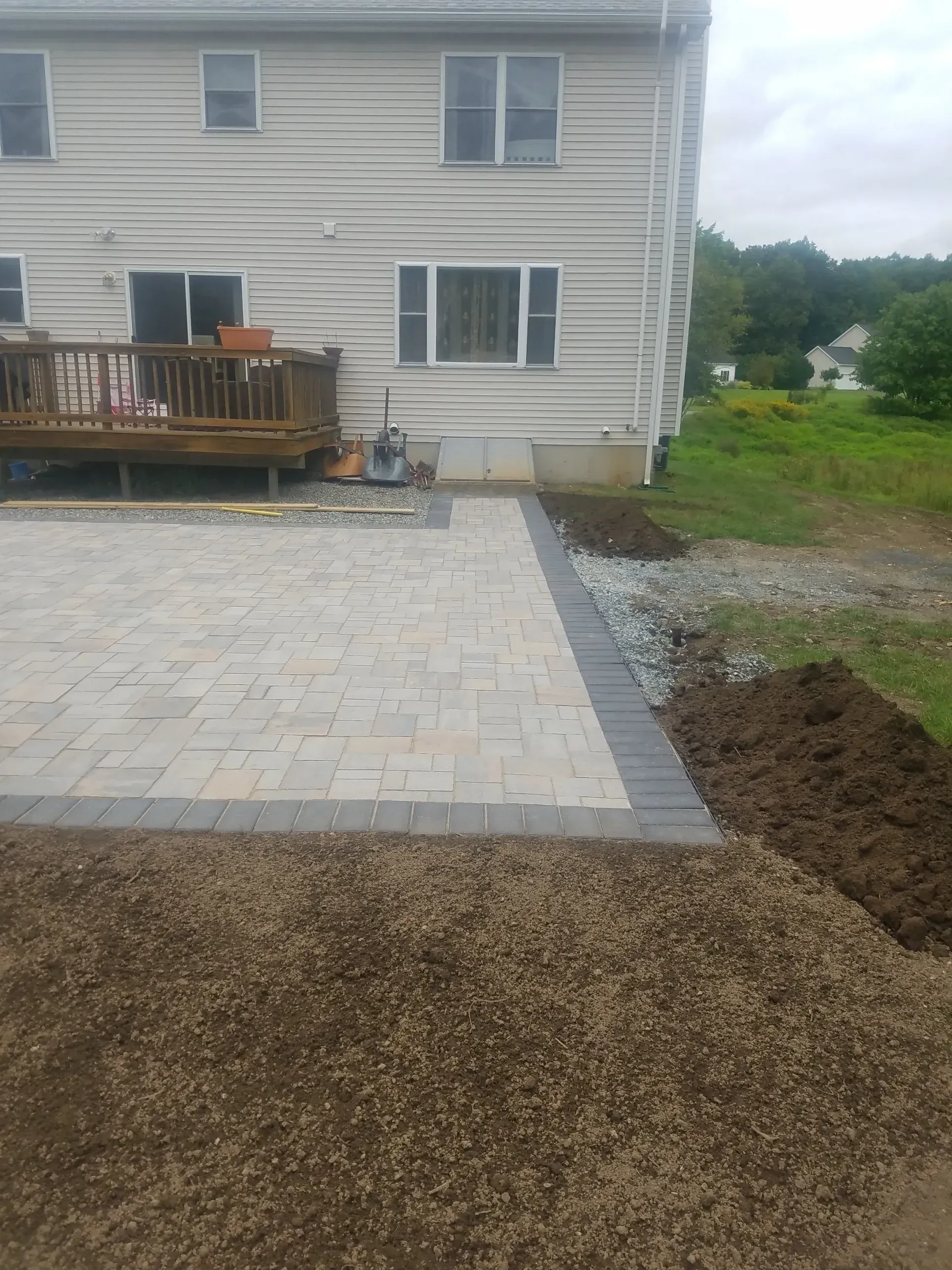 Backyard patio with gray pavers, wooden deck, and gravel path beside a house under construction