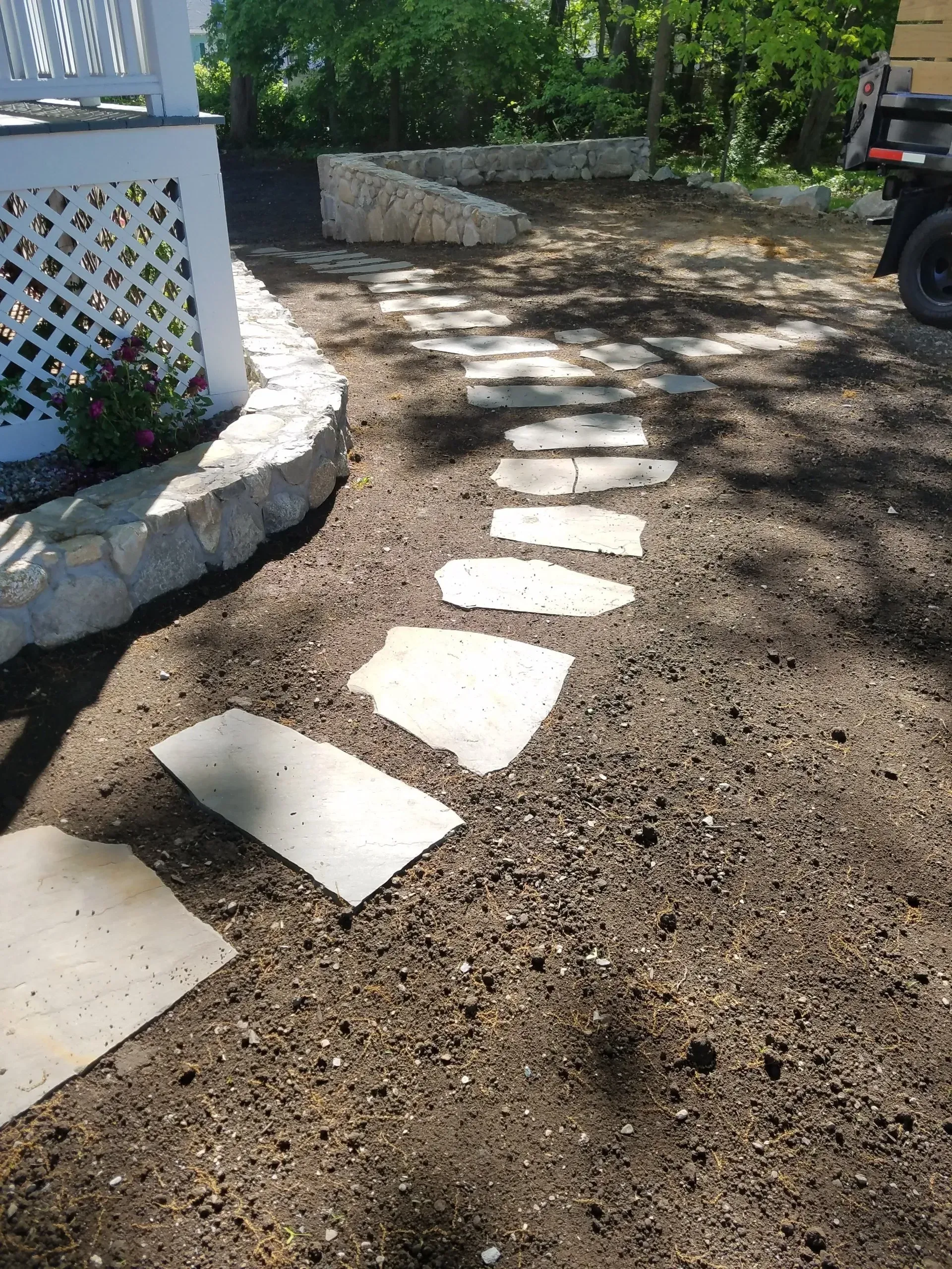 Curved stone stepping path through gravel beside a white lattice porch and garden, with sunlight and a parked vehicle.