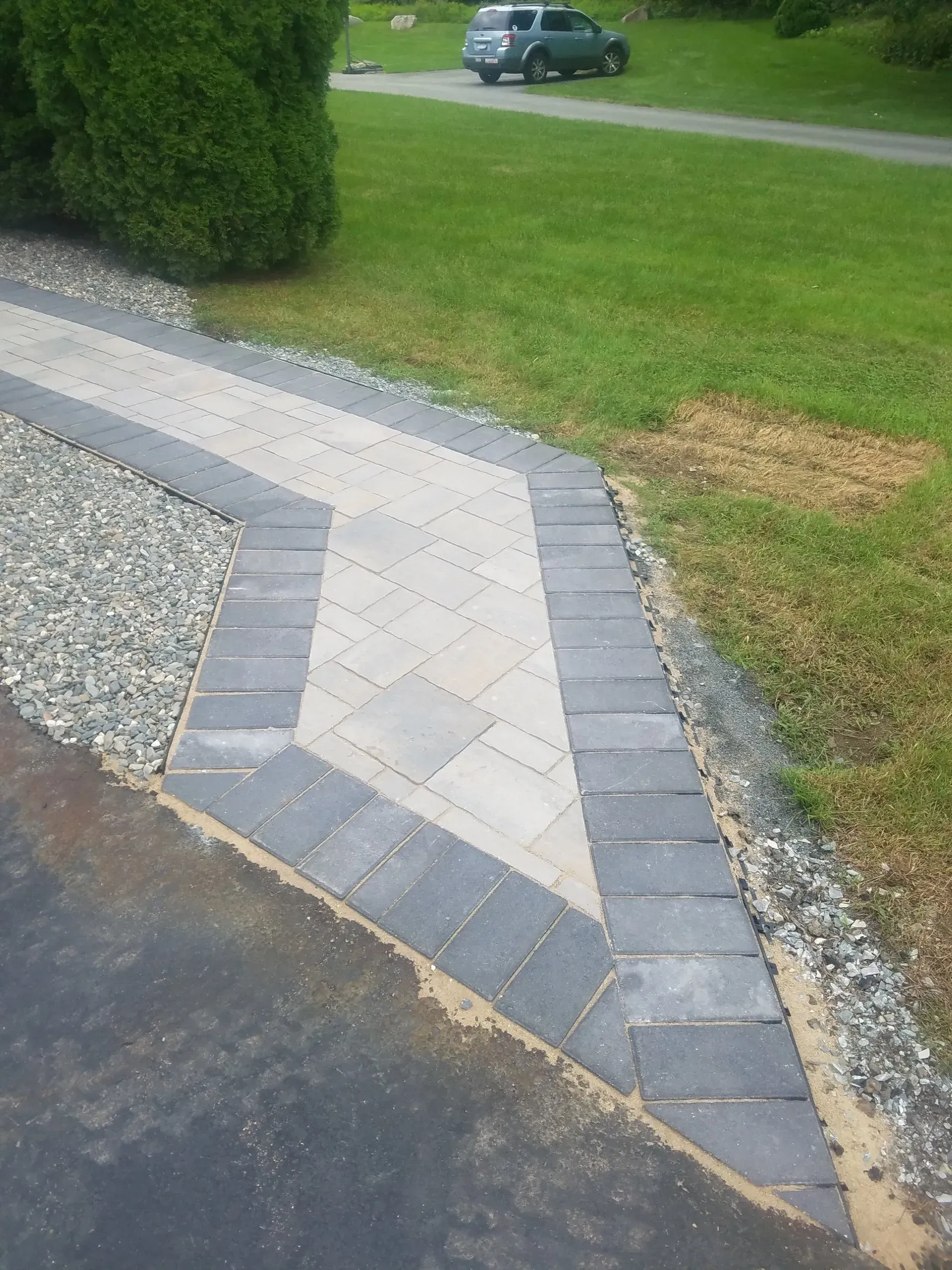 Concrete driveway edge with gray pavers beside grass and gravel, leading toward a parked car.