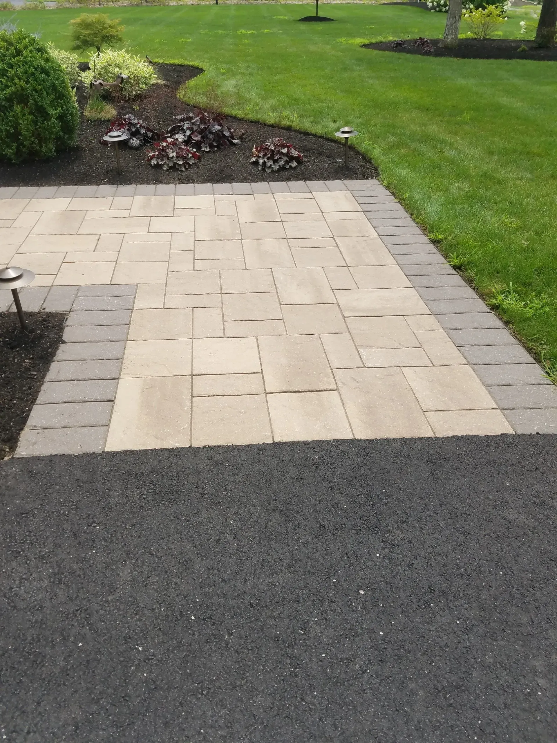 Paved walkway leading to a lawn and garden bed beside a dark asphalt surface.