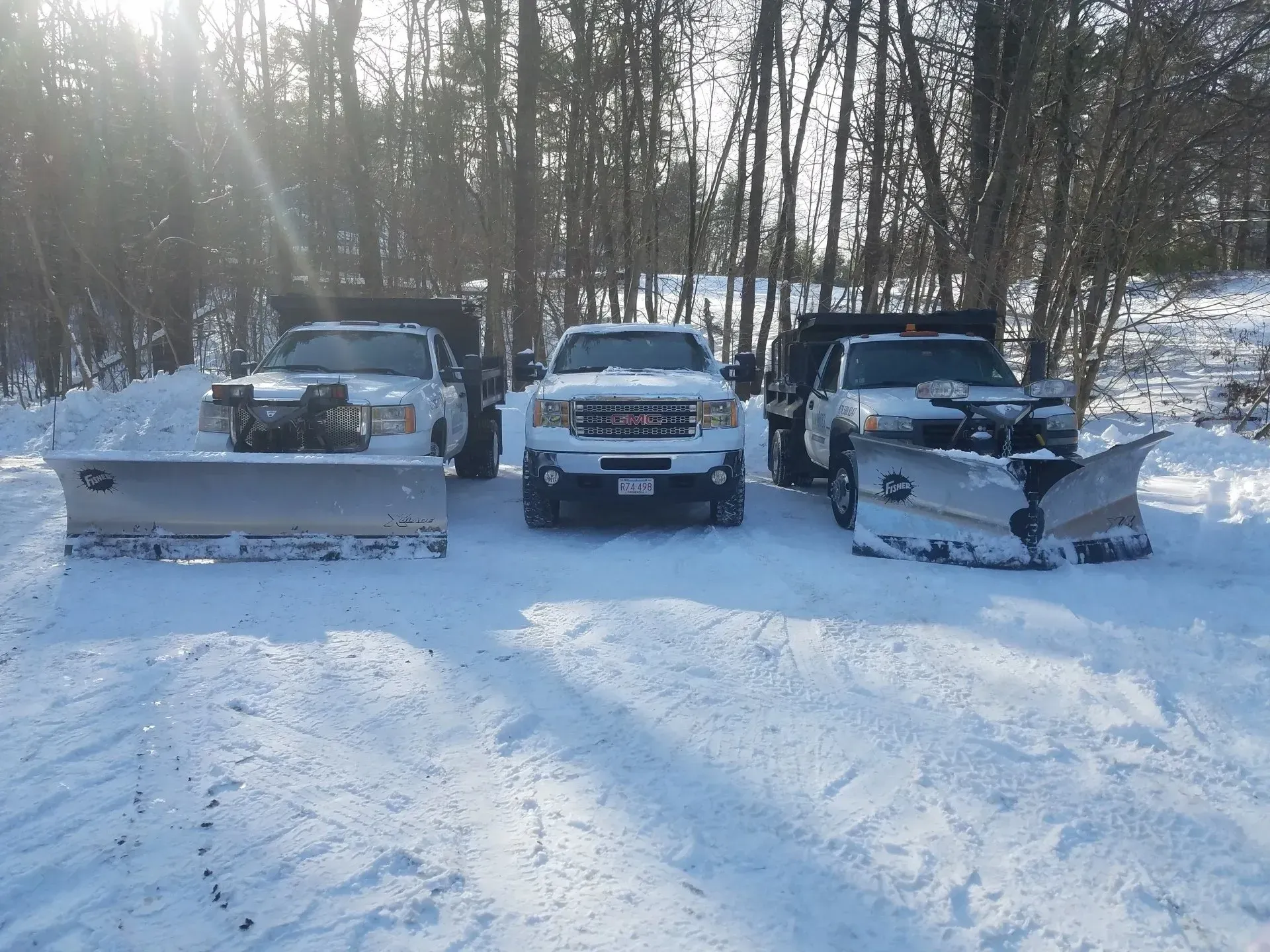 Three vehicles with snowplows parked in a snowy lot beside trees