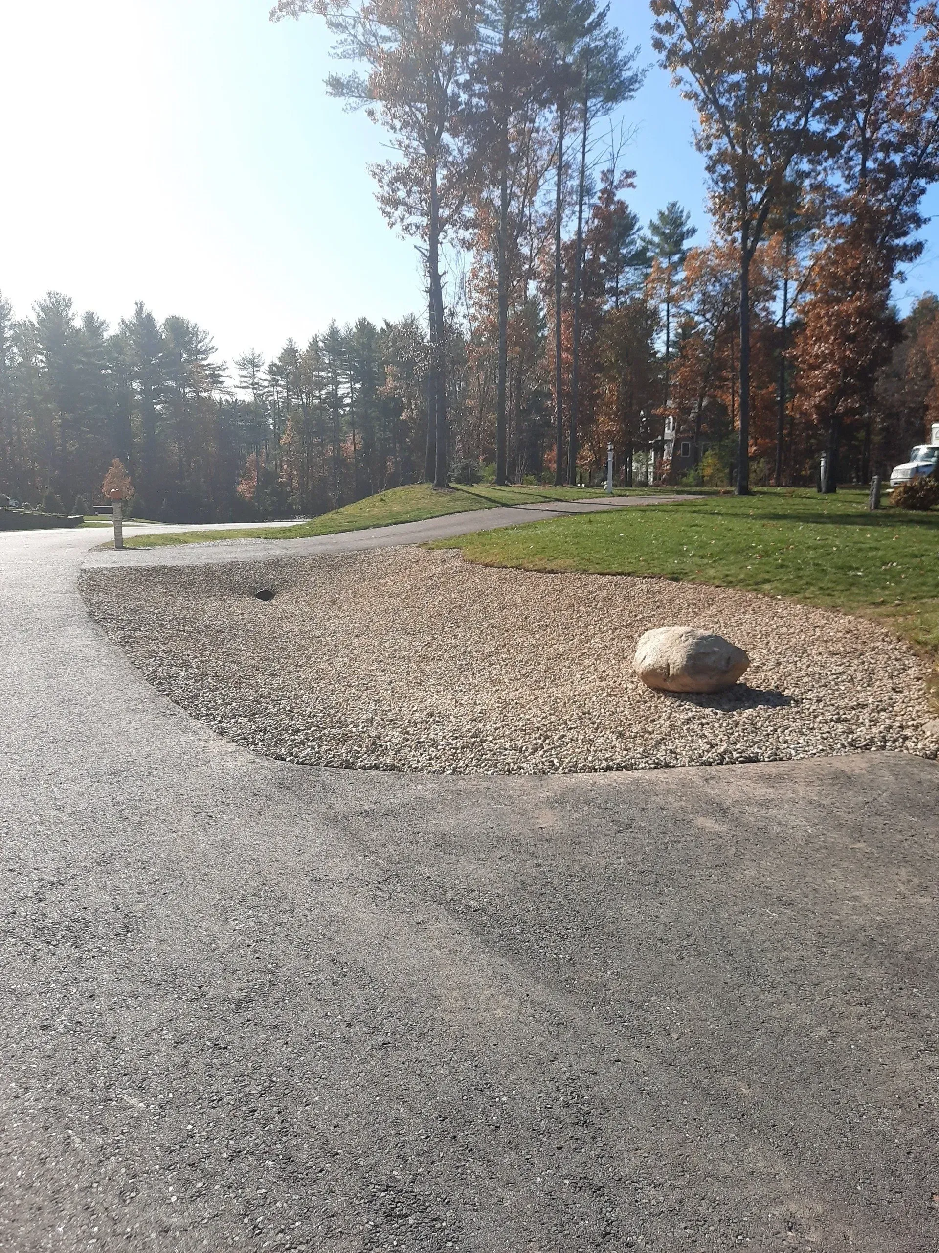Gravel parking area beside a grassy path and tall trees in bright sunlight, with a large rock in the foreground