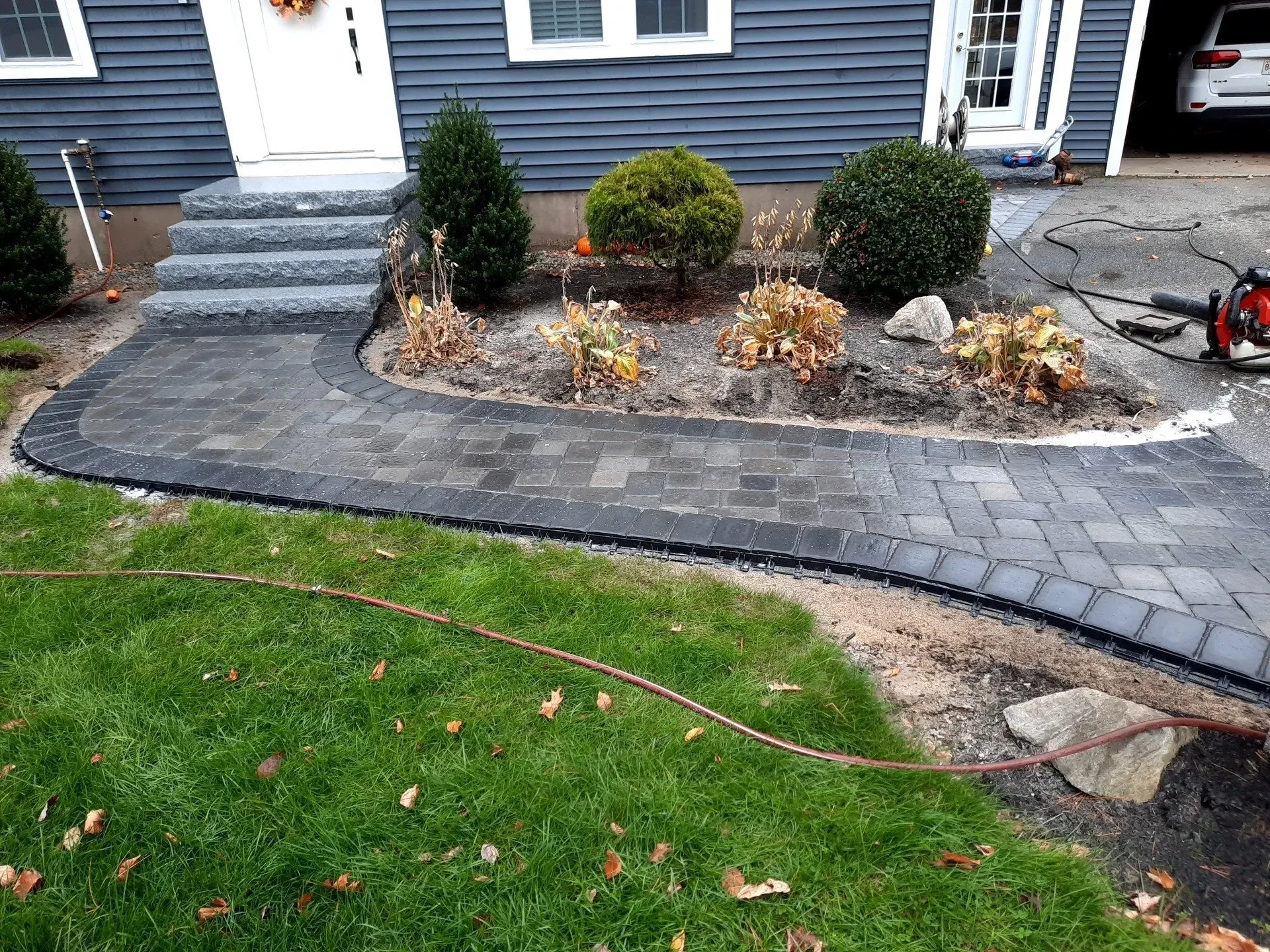 Front walkway with gray pavers, shrubs, and steps leading to a blue house; lawn in foreground.