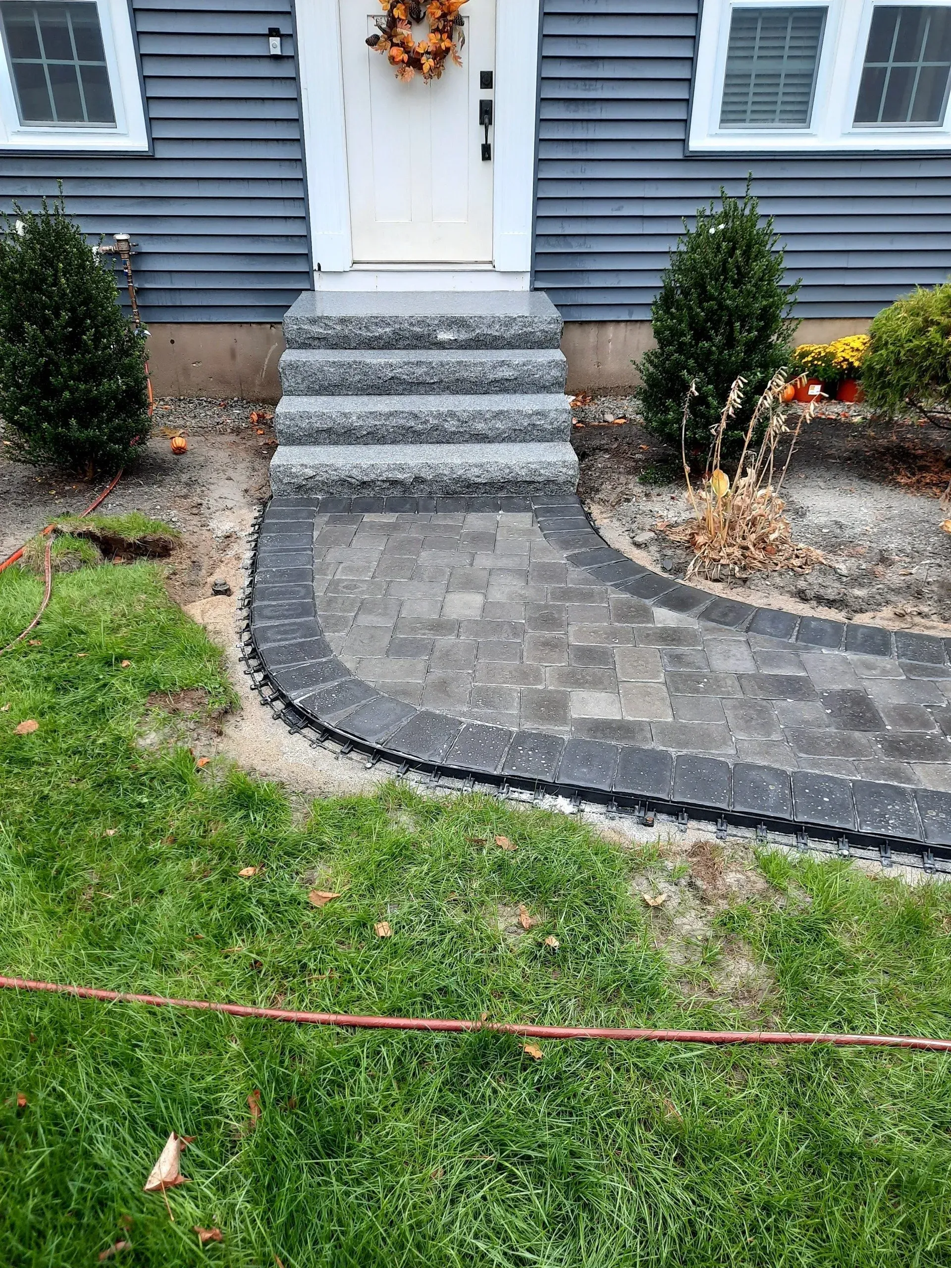 Front walkway of gray pavers leading to white front steps, bordered by black edging and shrubs.