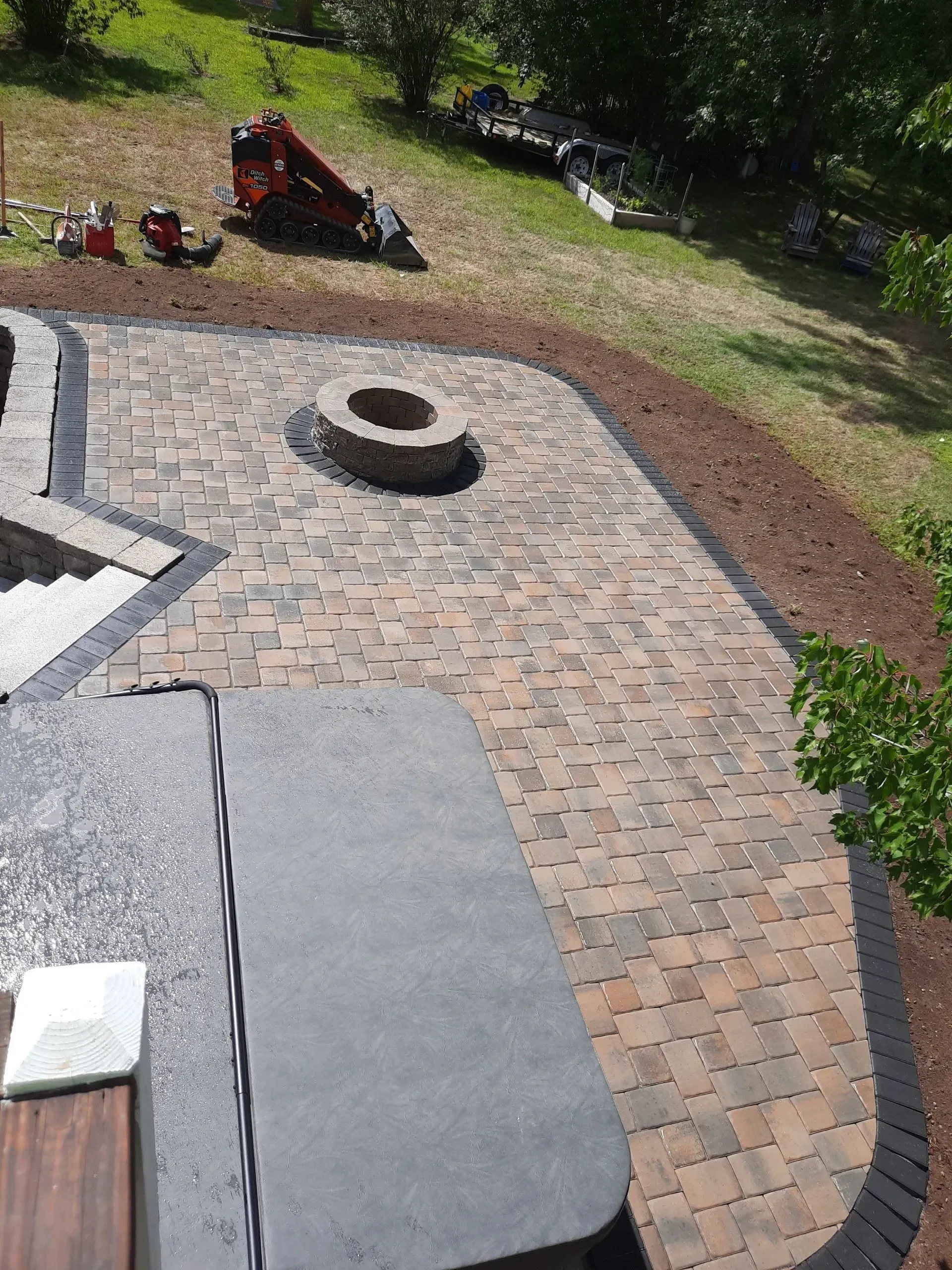 Backyard patio with brick pavers, a fire pit, and a red wheelbarrow near a grassy lawn.