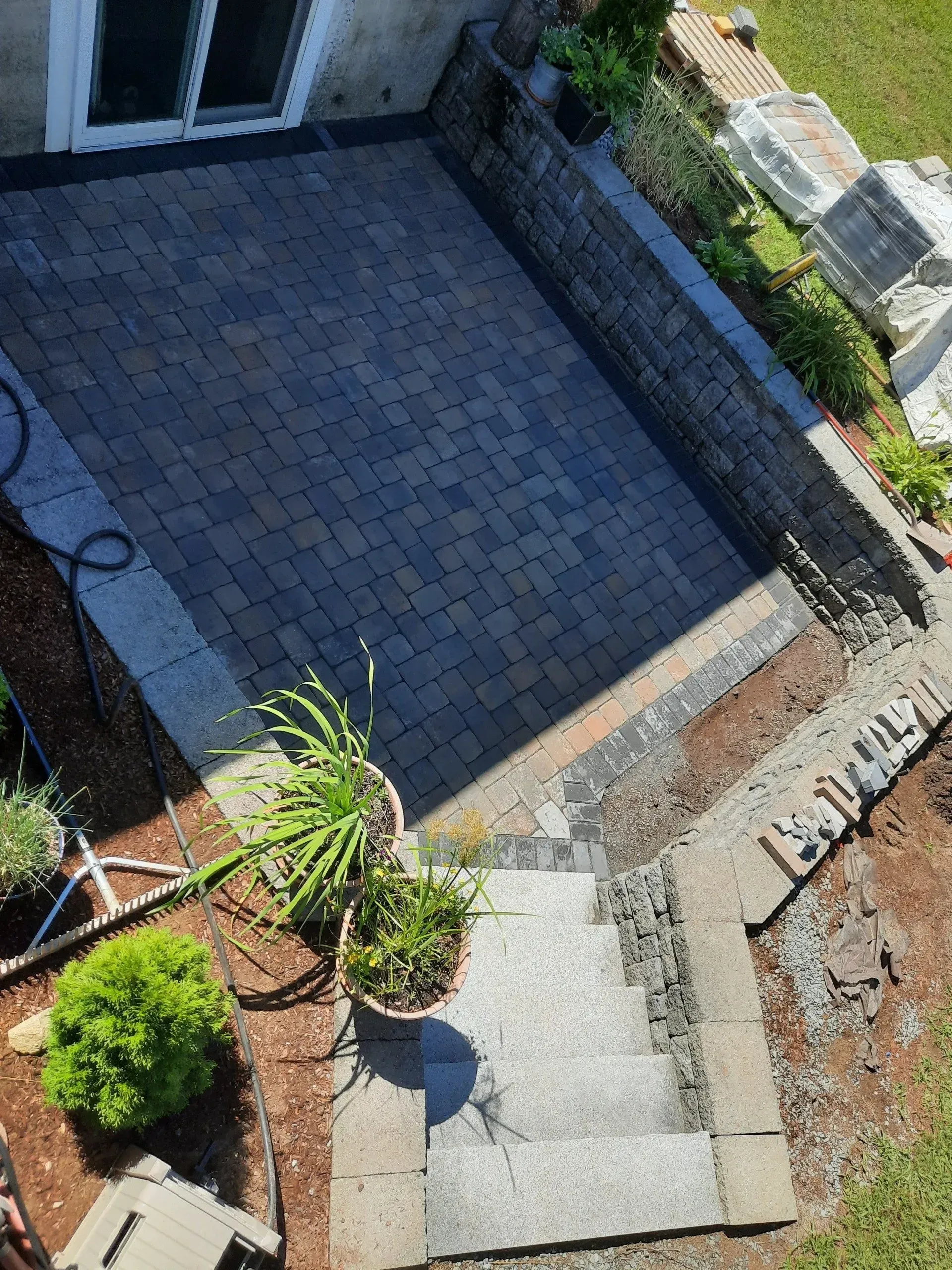 Small walled patio with gray pavers, steps, potted plants, and a hose beside a house entrance