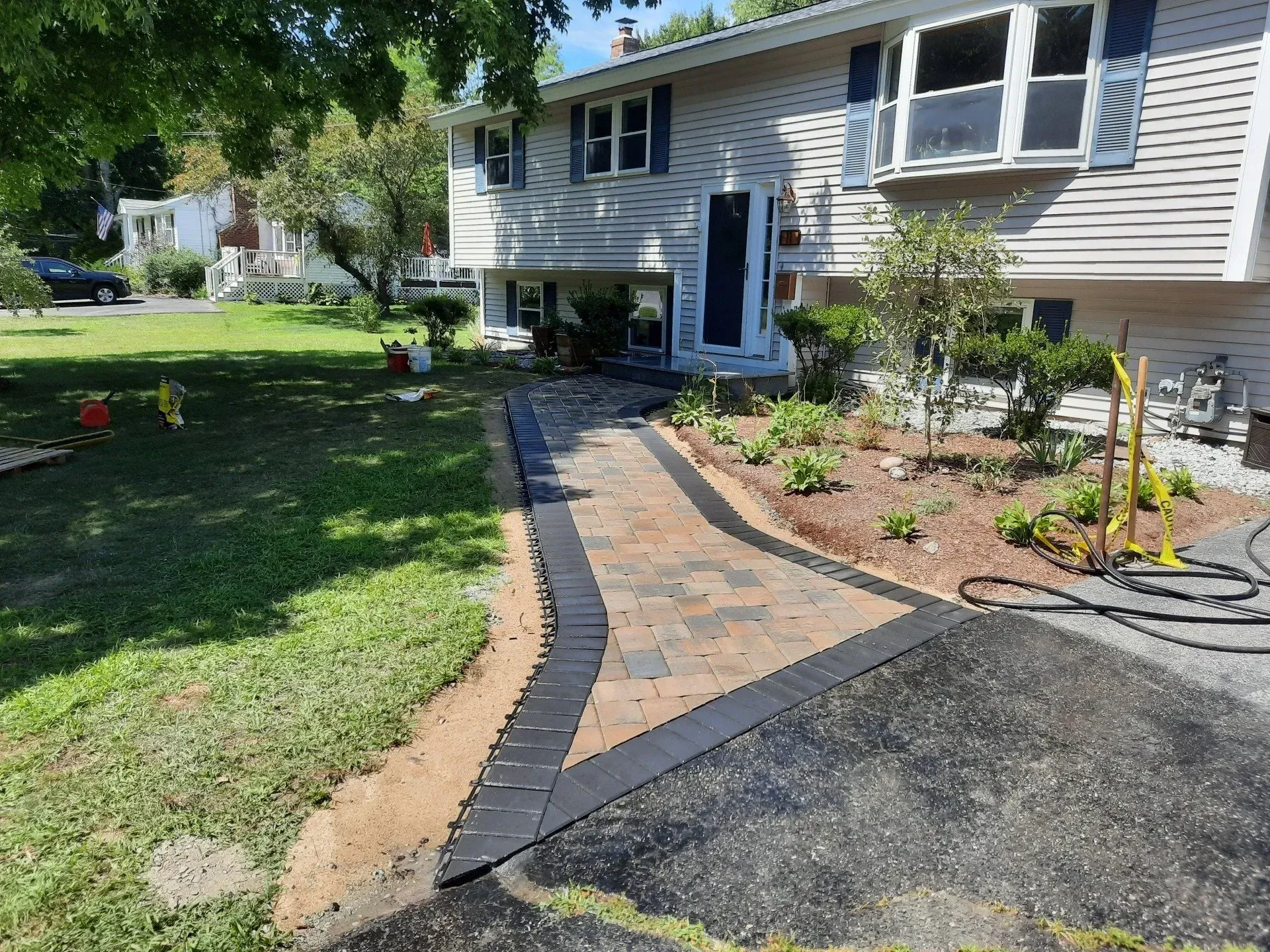 Front walkway with brick pavers and black edging leading to a gray house with a front door and garden beds.