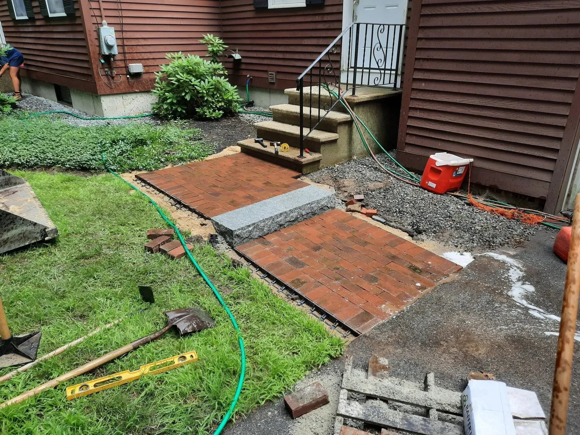 Backyard patio under construction with pavers, gravel, tools, and a hose beside a house