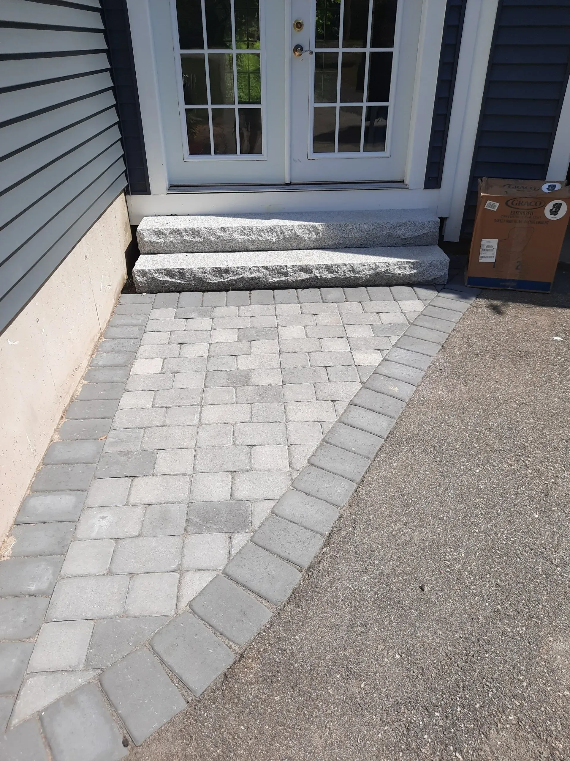 Front entry with paver walkway, stone steps, and double doors beside gray siding.