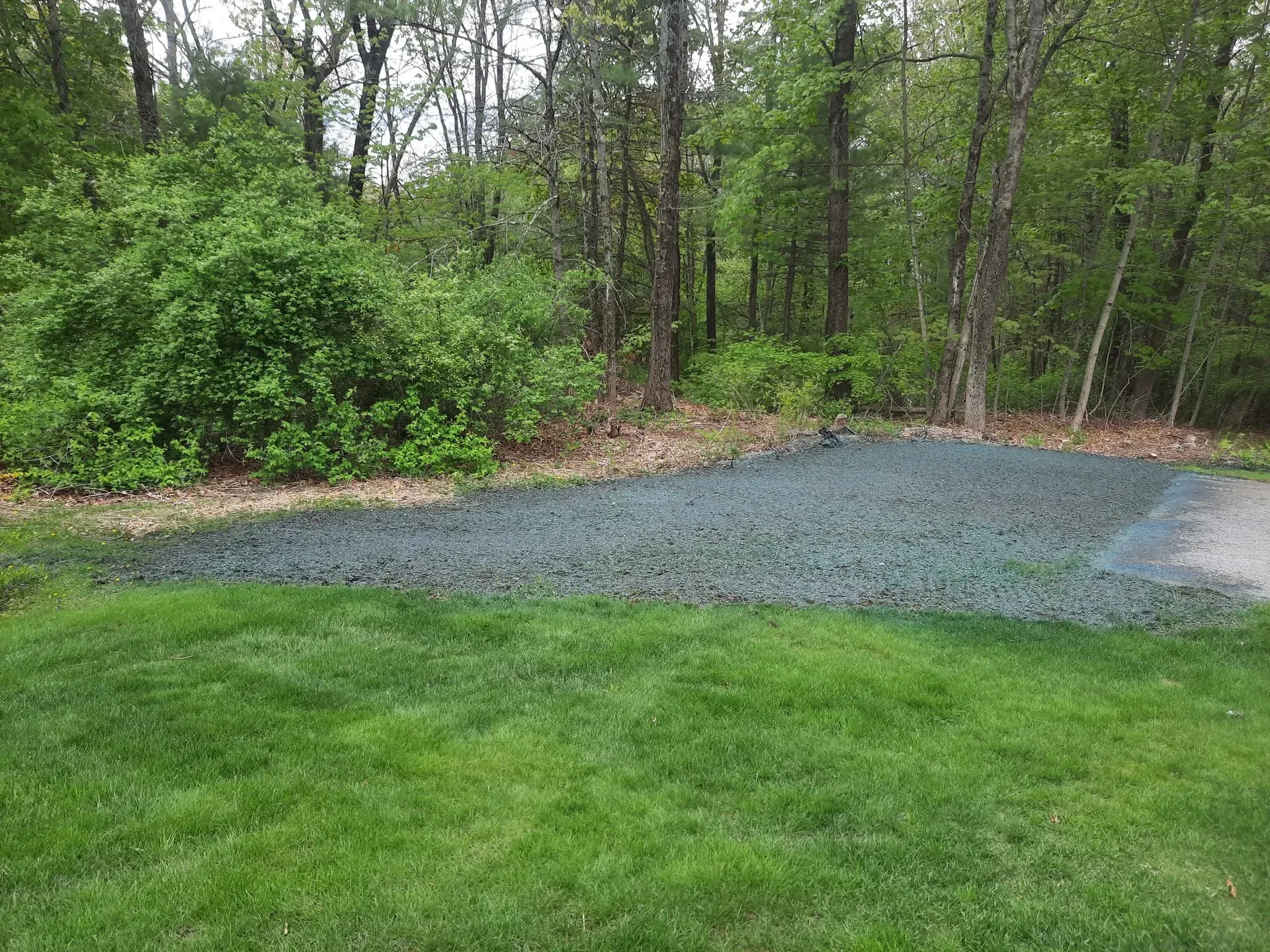 Grassy yard beside a gravel area with trees and shrubs in the background