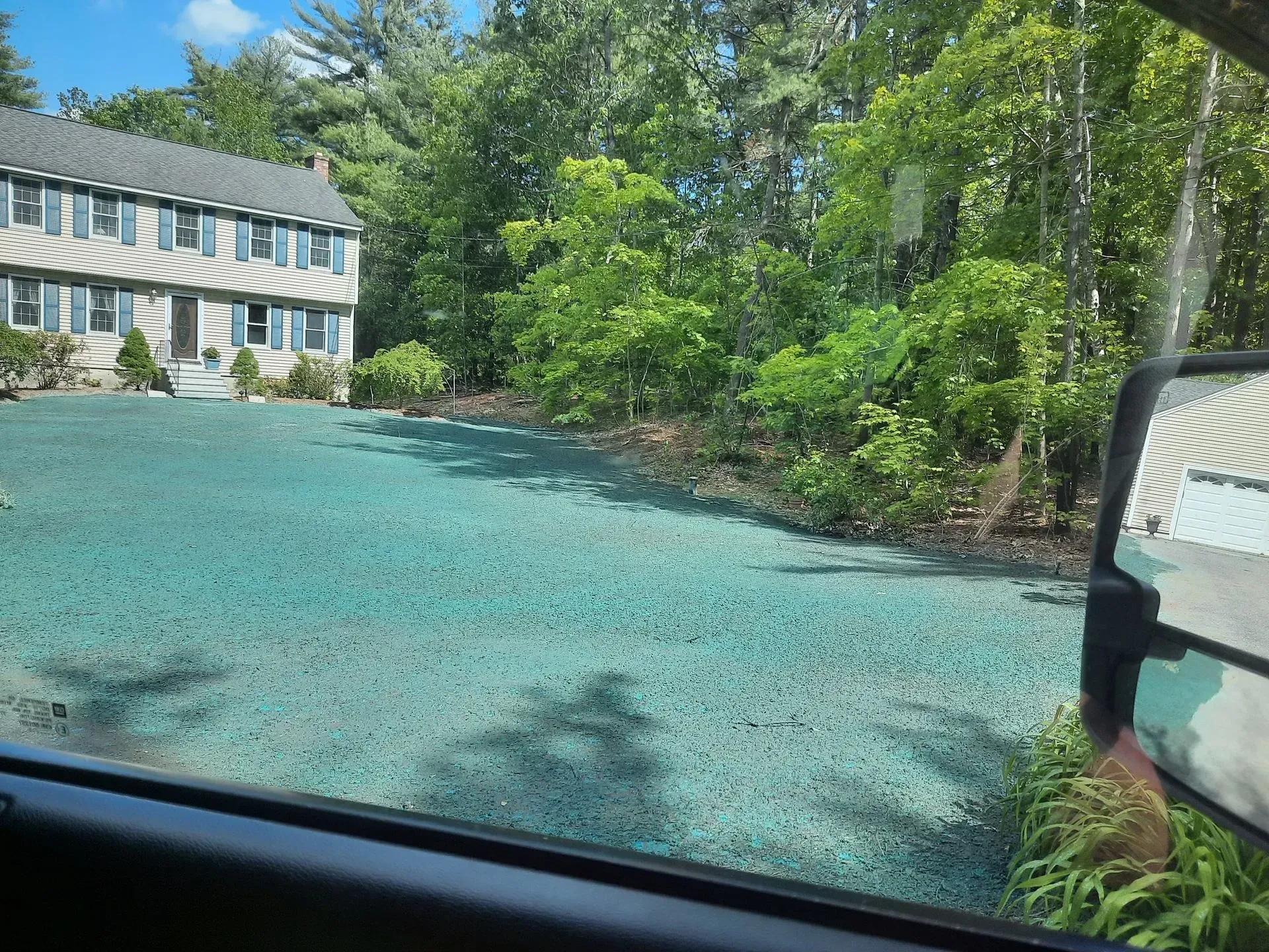 Gravel driveway beside a house and green trees, viewed from inside a vehicle.