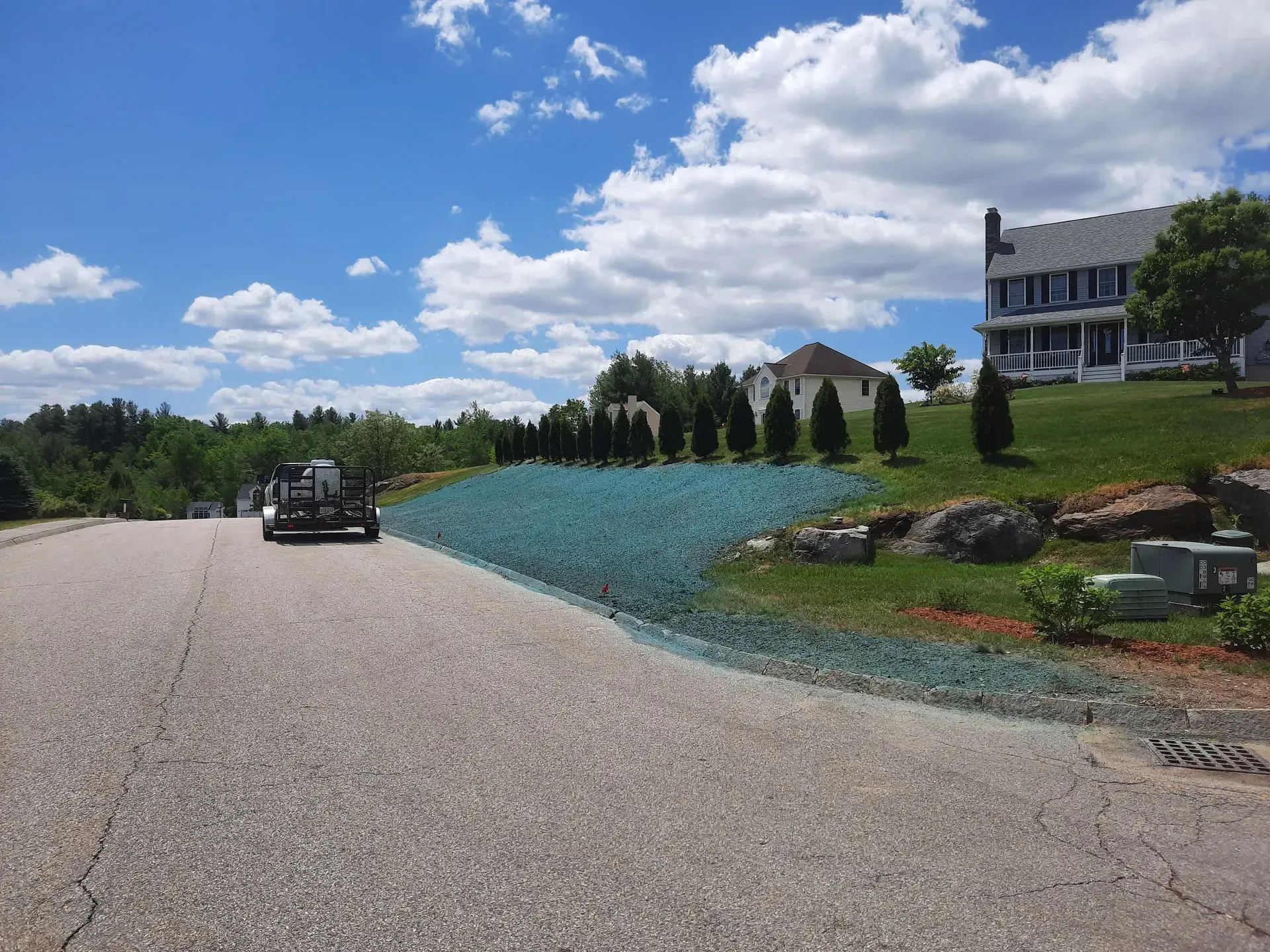 Curving gravel road beside blue flower bed, with a house and cloudy sky in the background.
