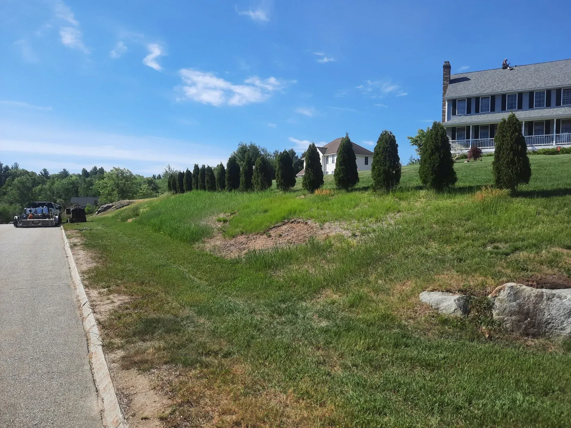 Grassy roadside beside a white building and evergreens under a blue sky; parked cars line the road.